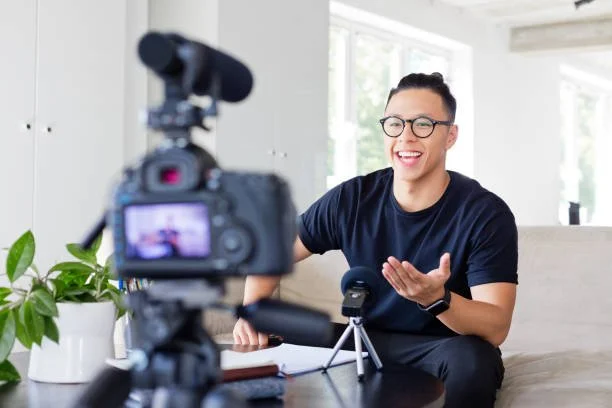 Man with glasses smiling and speaking into a microphone during a video recording or interview at home, with camera and recording equipment on a table