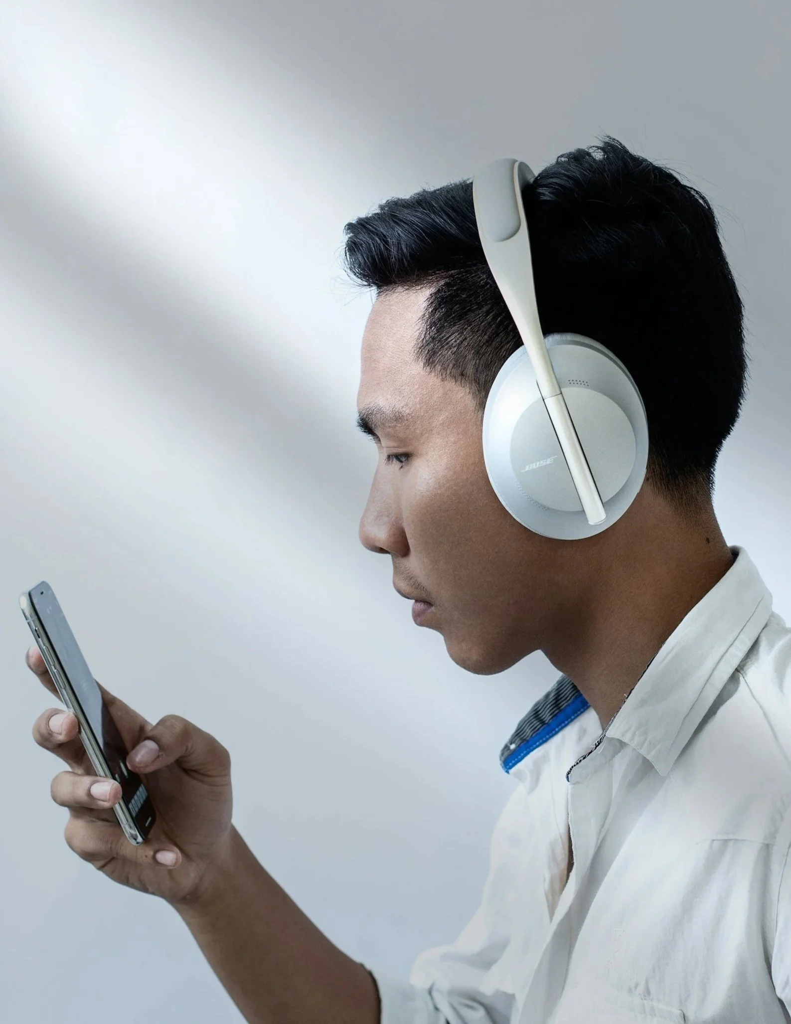 A young man wearing white Bose headphones and a white shirt, looking at his smartphone against a light gray background.