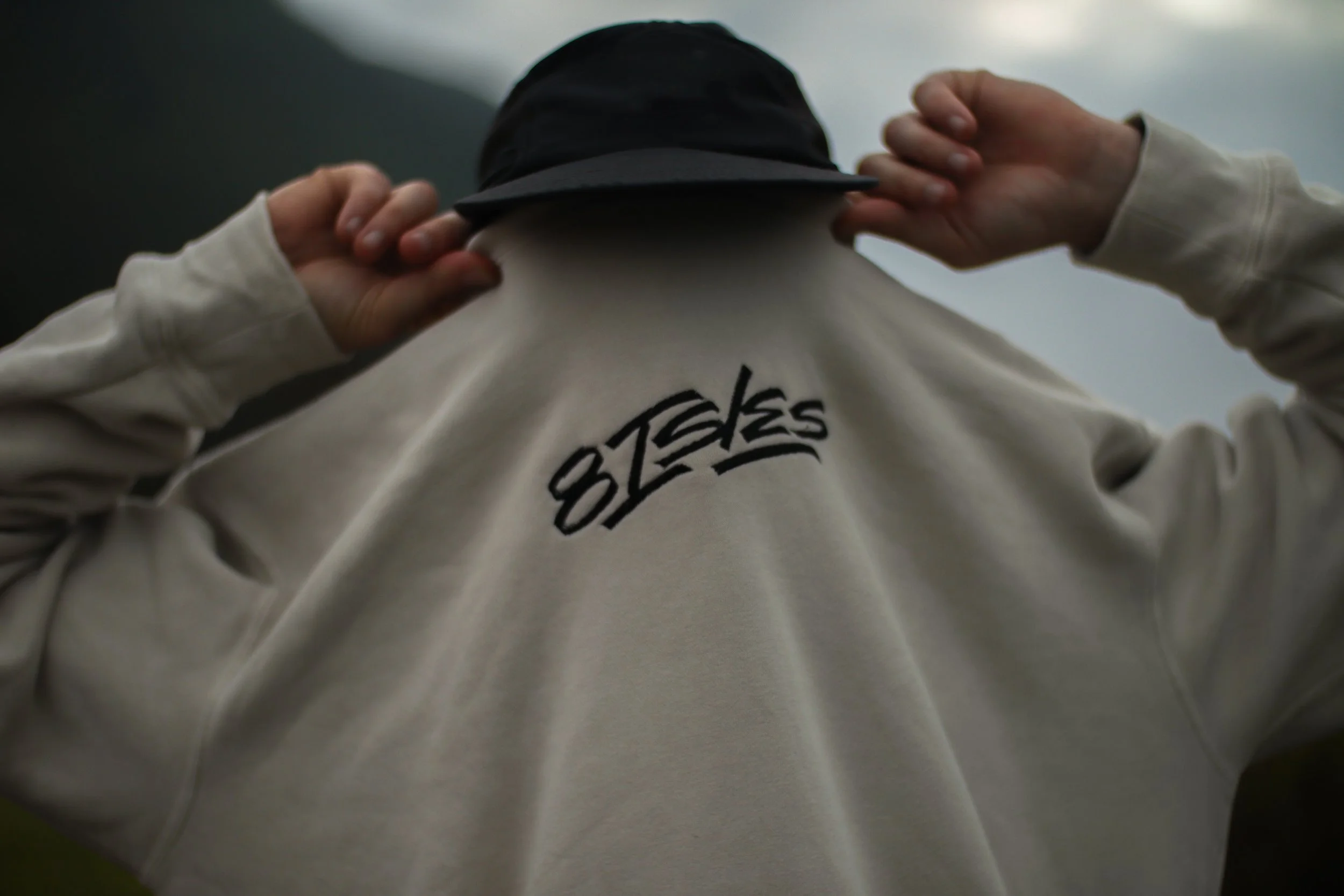 Person adjusting a black cap, wearing a cream hoodie with the word 'STEPS' embroidered on the back, standing outdoors with a cloudy sky in the background.