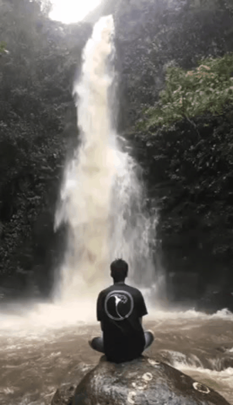 Person sitting cross-legged on a rock facing a tall waterfall in a forested area.