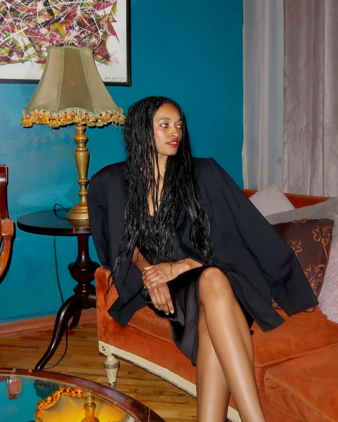 A woman with long curly hair sitting on an orange velvet couch in a living room, with a gold lamp, blue wall, and a colorful abstract painting on the wall behind her.