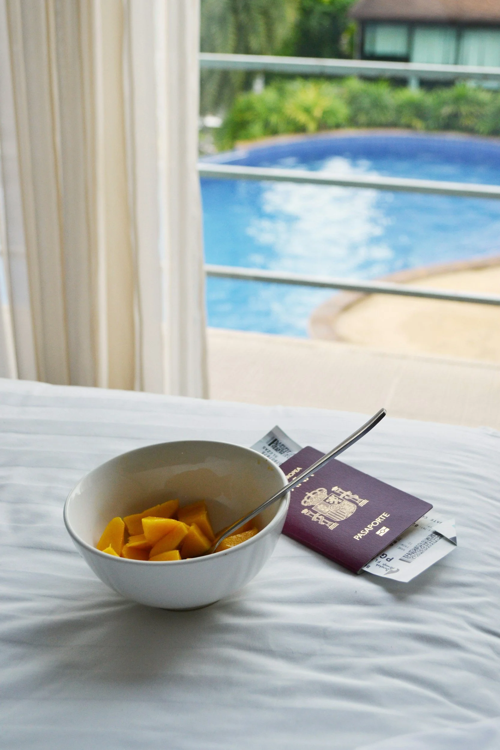 A white bowl of sliced mango on a bed with a spoon, a Paraguayan passport, and a boarding pass, with a view of a swimming pool and greenery through a window.