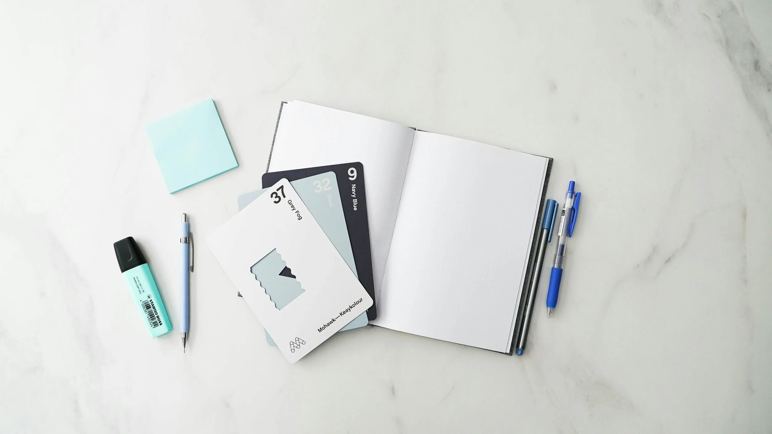 Open notebook with pens, stationery, paint palette featuring grey fog, navy blue, and black paint colors, sticky notes, and paint swatch cards on a white marble surface.