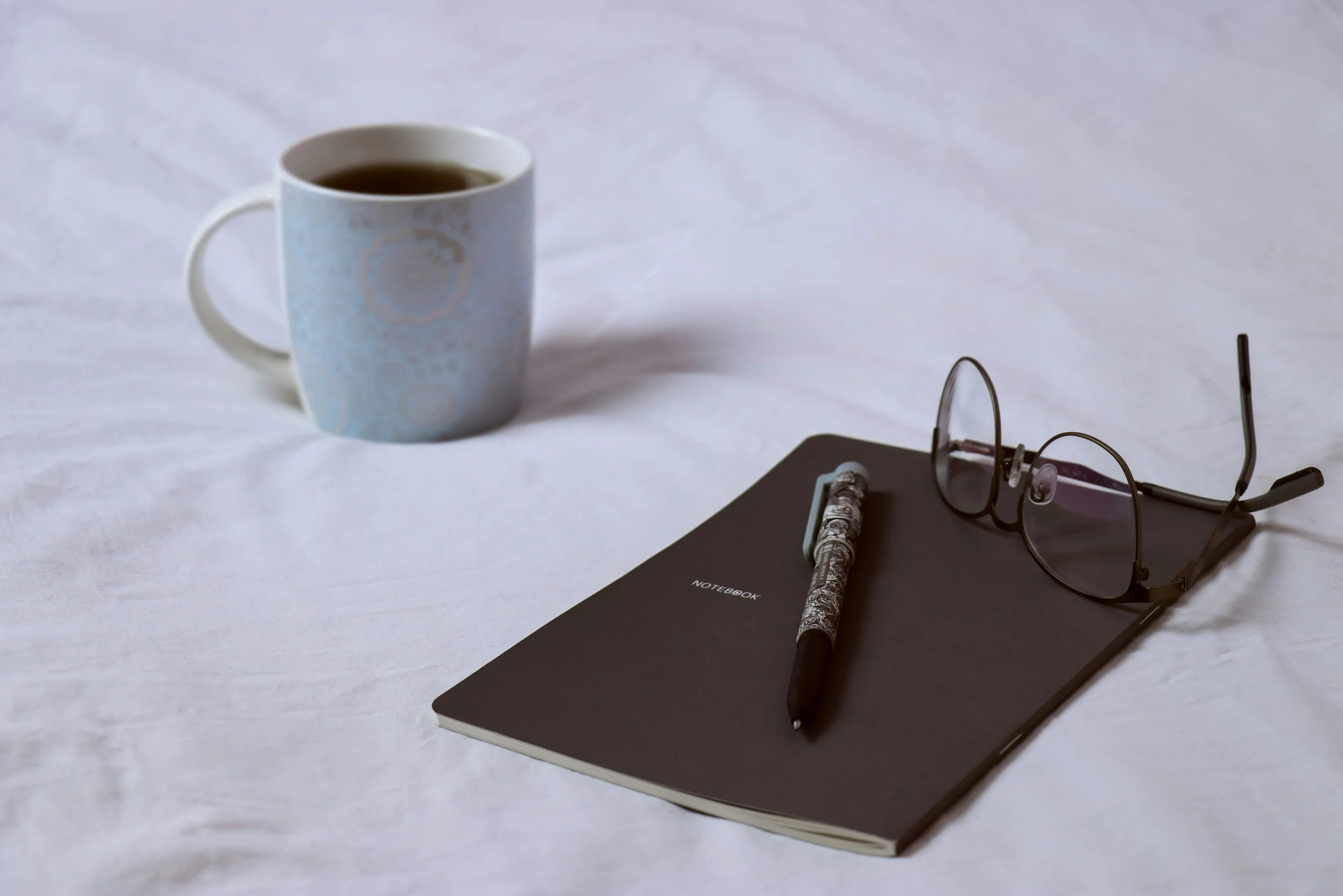 A white coffee mug filled with black coffee, a gray notebook labeled 'NOTEBOOK', a black and white patterned pen, and a pair of black-rimmed glasses resting on the notebook, all on a white surface.