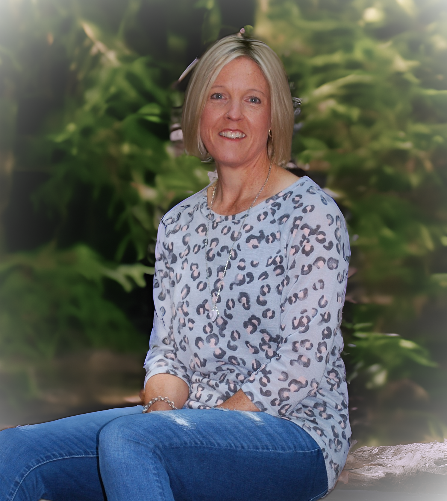 A woman with blonde hair wearing a leopard print shirt sitting outdoors in front of green foliage.