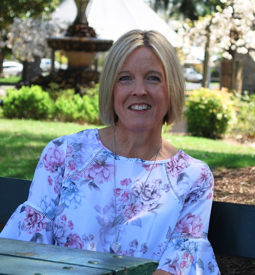 A smiling woman with blonde hair and a floral blouse sitting outdoors on a park bench, with trees, greenery, and a fountain in the background.