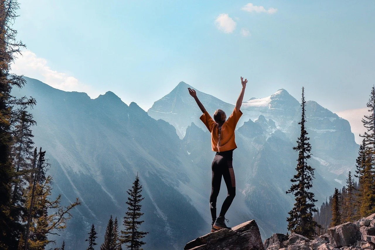 A person standing on a rock with arms raised, facing mountains and a blue sky.