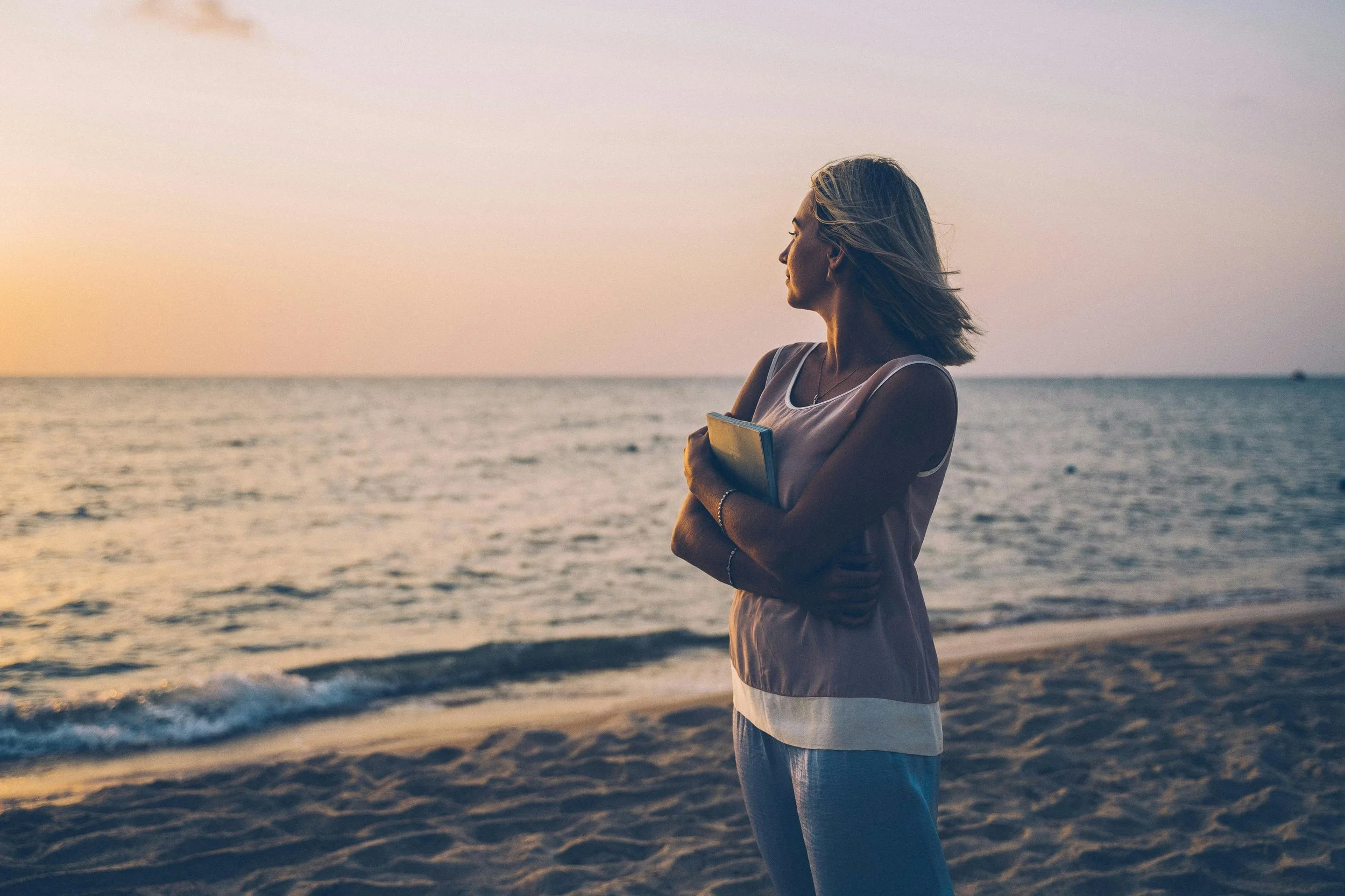 Woman standing on the beach holding a notebook, gazing at the ocean at sunset.