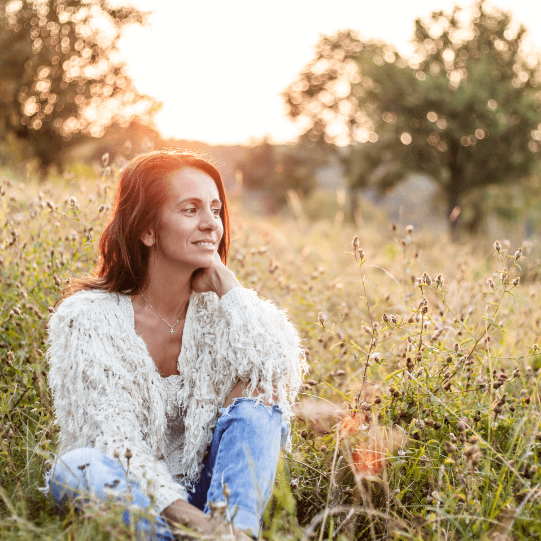 A woman with red hair sitting in a field with wildflowers, illuminated by sunset sunlight.