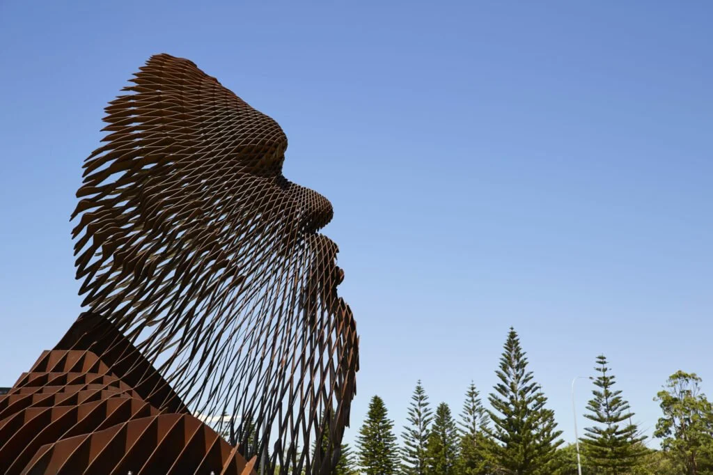 Large sculpture of a person's profile made of layered, rust-colored metal pieces, with trees and a clear blue sky in the background.