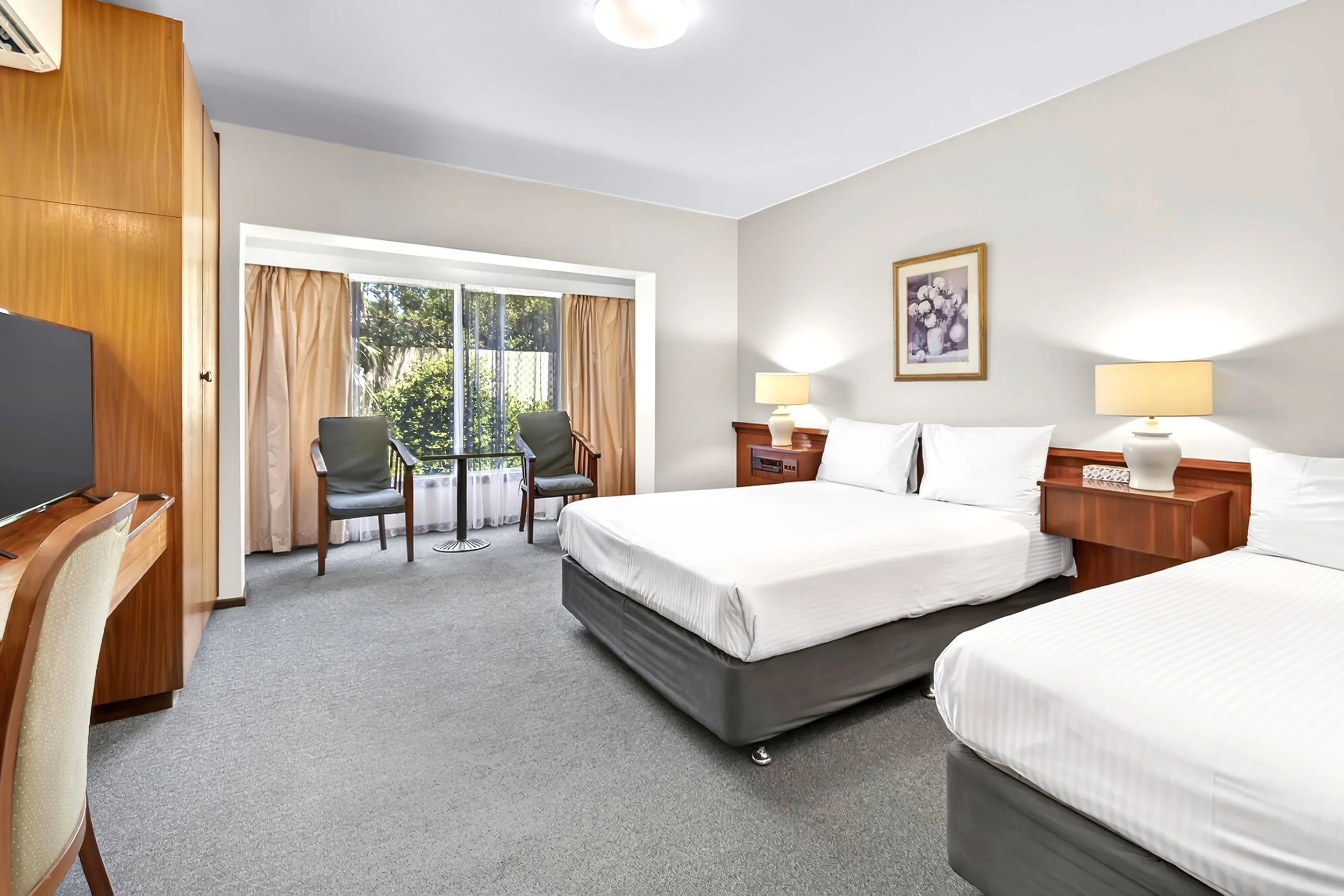 A hotel room with two beds, a small sitting area near a large window, and a television on a wooden desk.