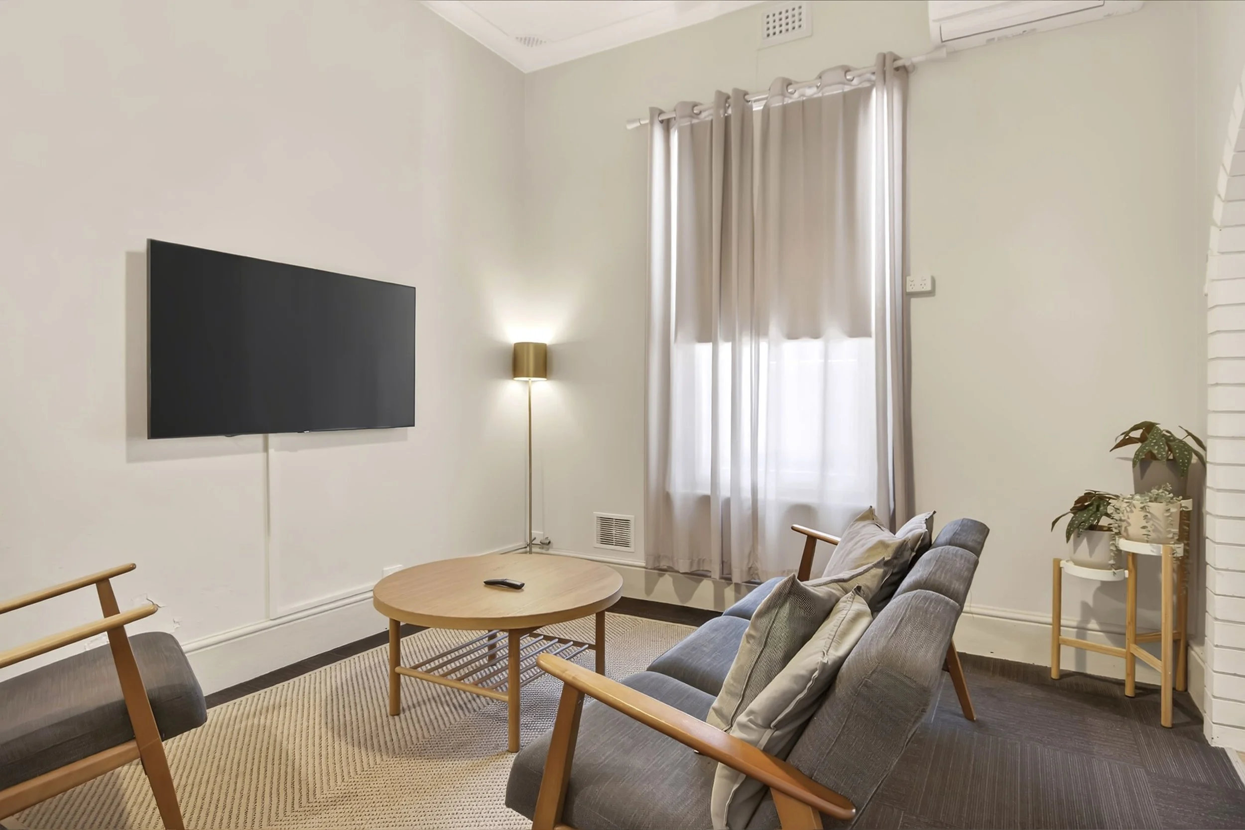 Living room with a wall-mounted flat-screen TV, a side standing lamp, beige curtains covering a window, a gray sofa with throw pillows, a circular wooden coffee table with a remote, and potted plants on a wooden stand.