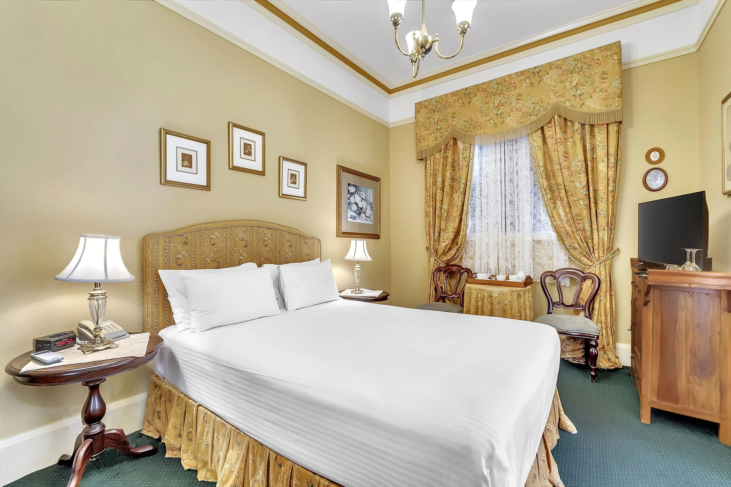 A hotel room with a bed, bedside lamps, chairs, a window with curtains, a dresser with a TV, and framed artwork on the wall.