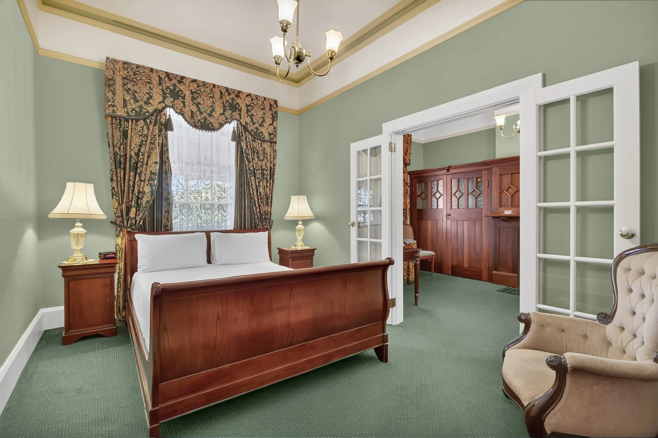 A vintage-style bedroom with green walls, a wooden bed with white bedding, two bedside lamps, a lace-curtained window, a beige upholstered armchair, and a doorway leading to a closet area with wooden paneling.