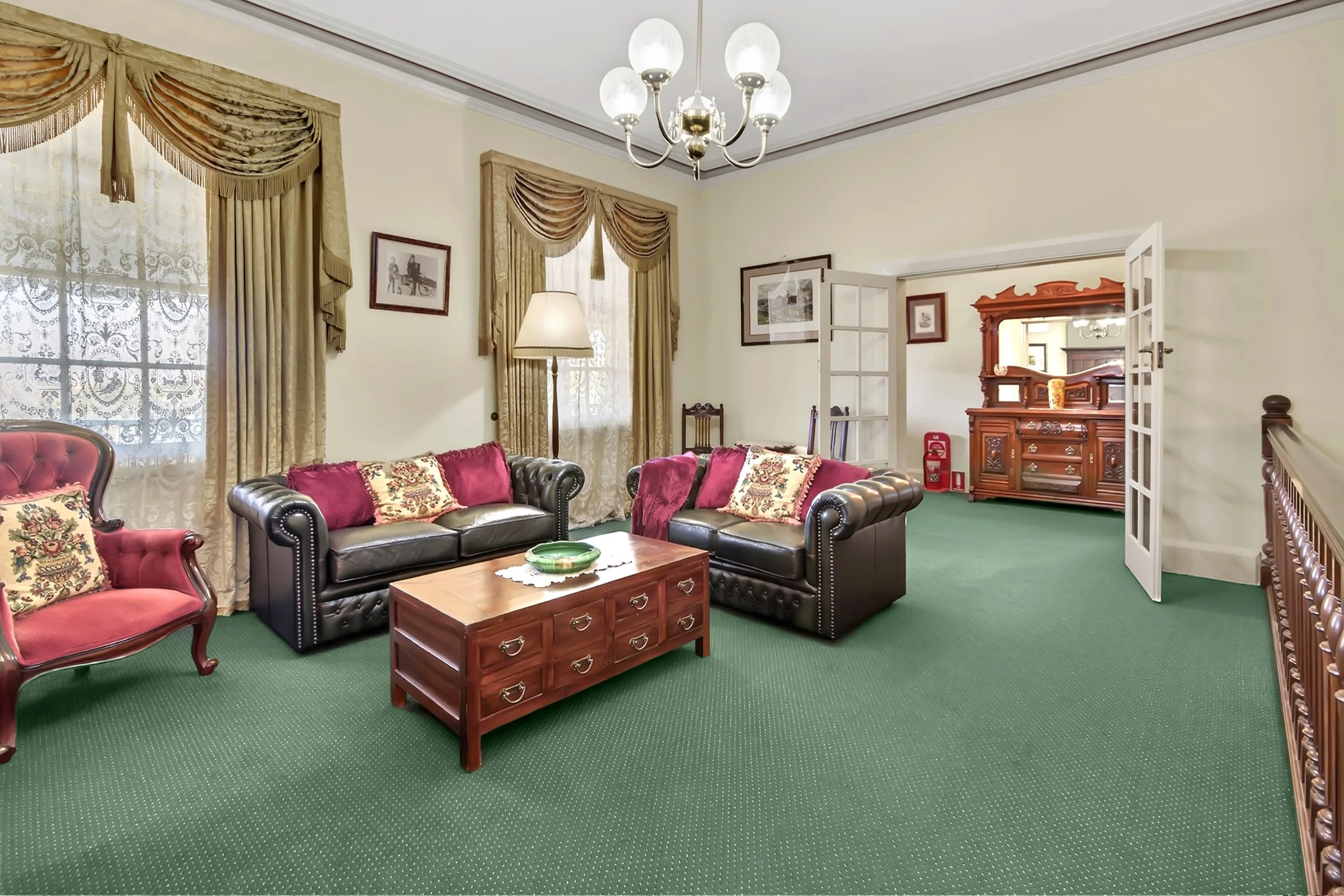 Living room with dark brown leather sofas, pink and floral throw pillows, wooden coffee table, vintage-style curtains, chandelier, and an open doorway leading to another room with a wooden sideboard and mirror.