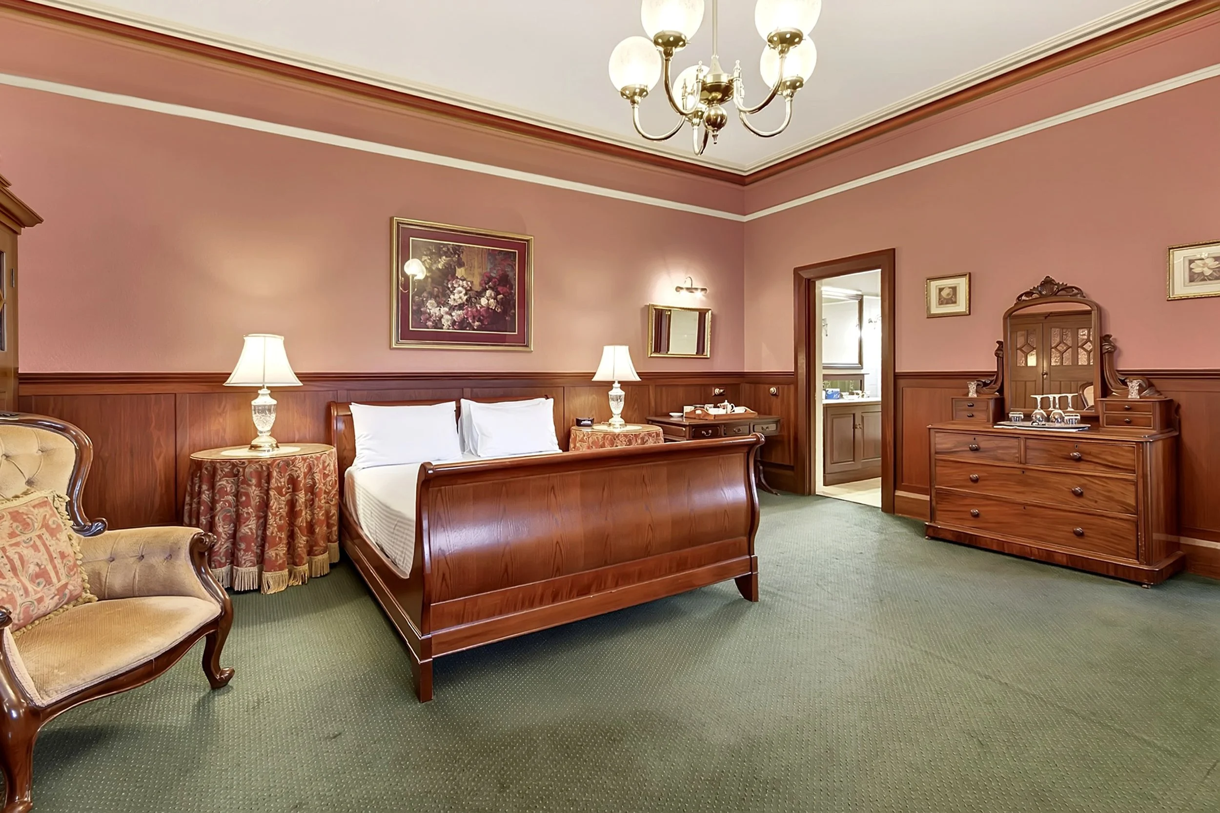 A hotel room with a wooden bed, two lamps, a wooden dresser with a mirror, a vintage armchair, wall art, and a doorway leading to the bathroom.