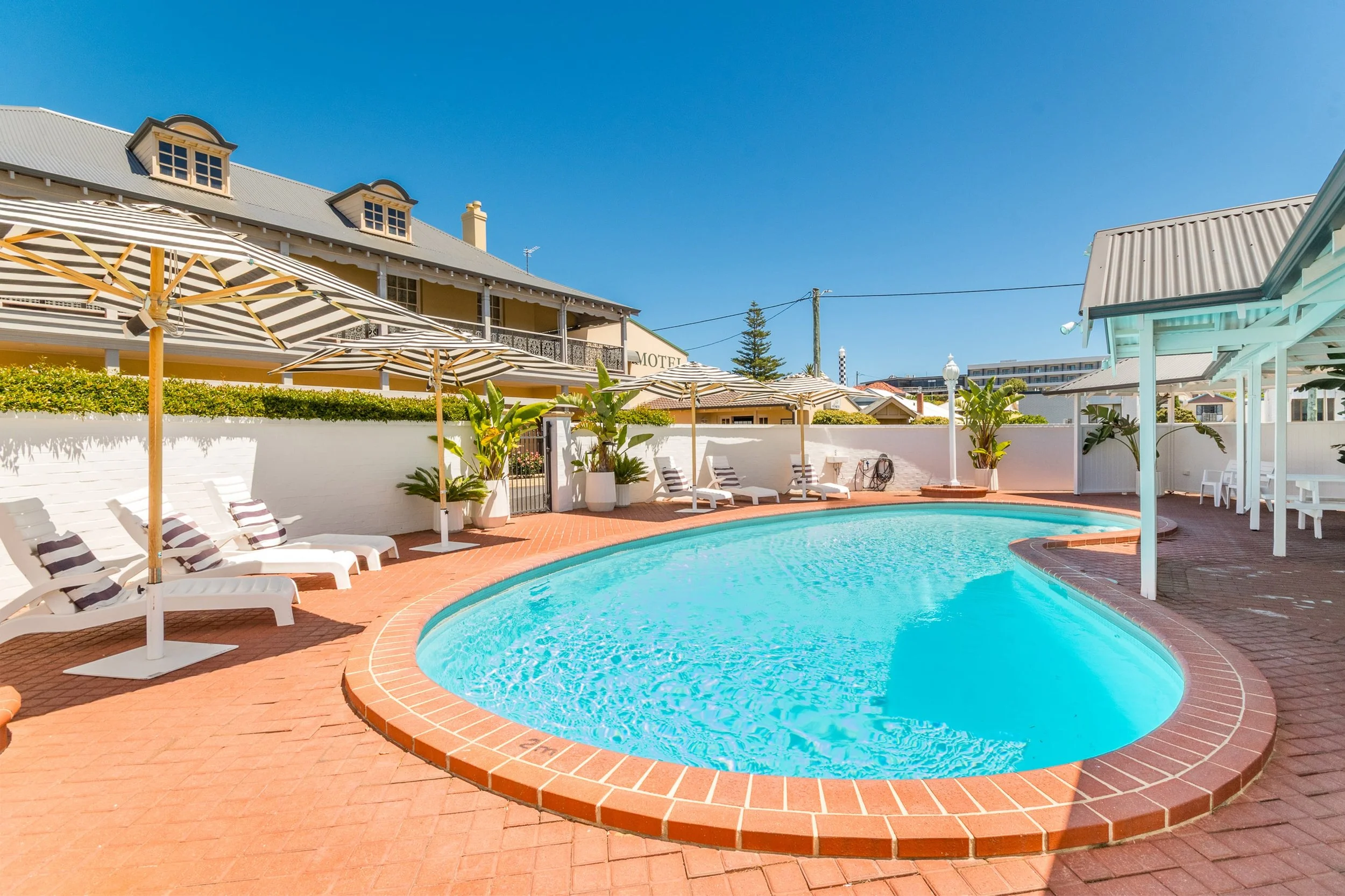 A hotel courtyard with a kidney-shaped swimming pool, multiple white lounge chairs with striped cushions, and striped umbrellas. Potted plants and a white fence surround the area. A building with a motel sign is in the background under a clear blue s