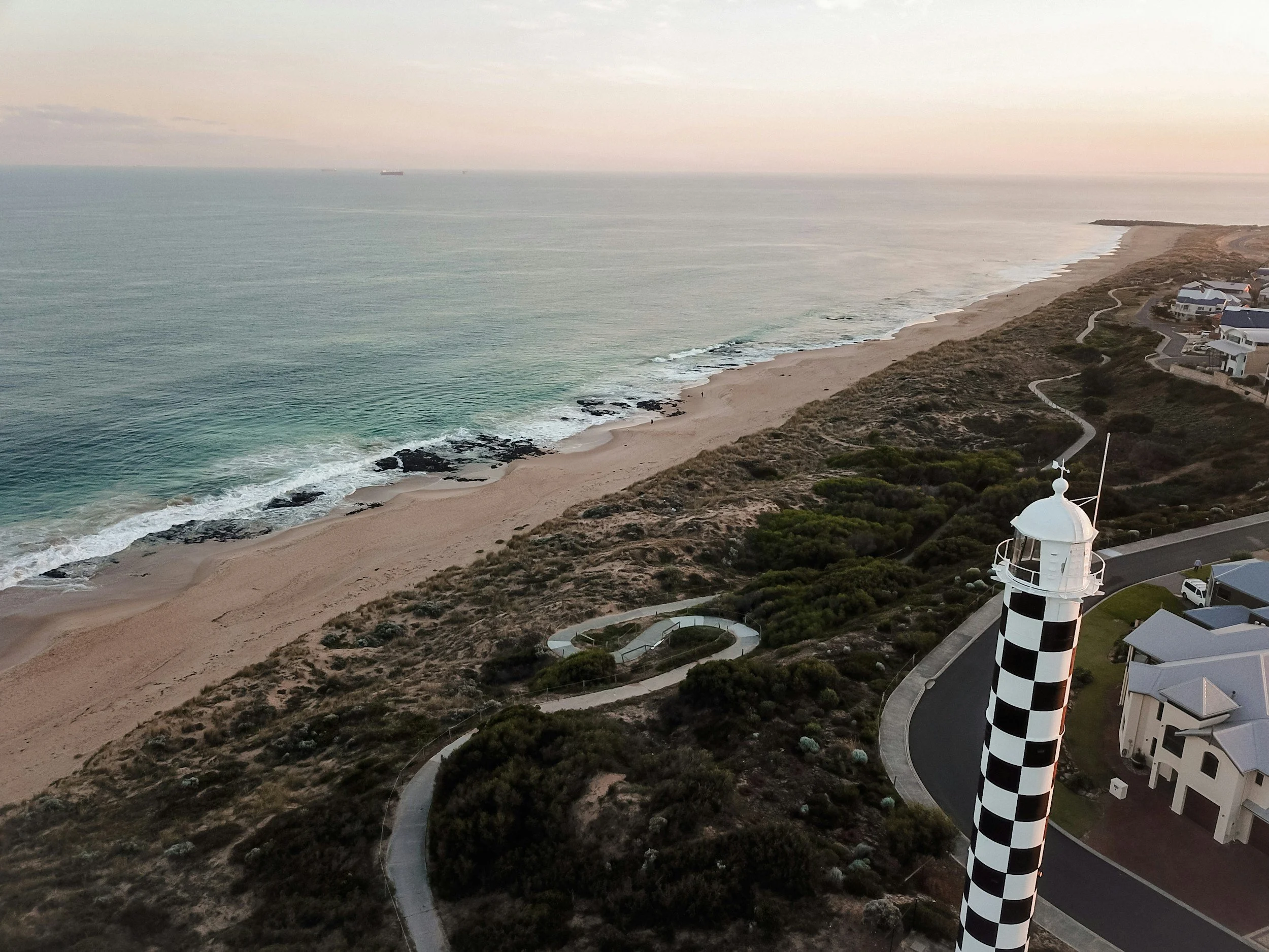 A coastal landscape showing a sandy beach, ocean waves, a lighthouse with black and white checkered pattern, a winding road, and some houses along the shoreline during sunset.