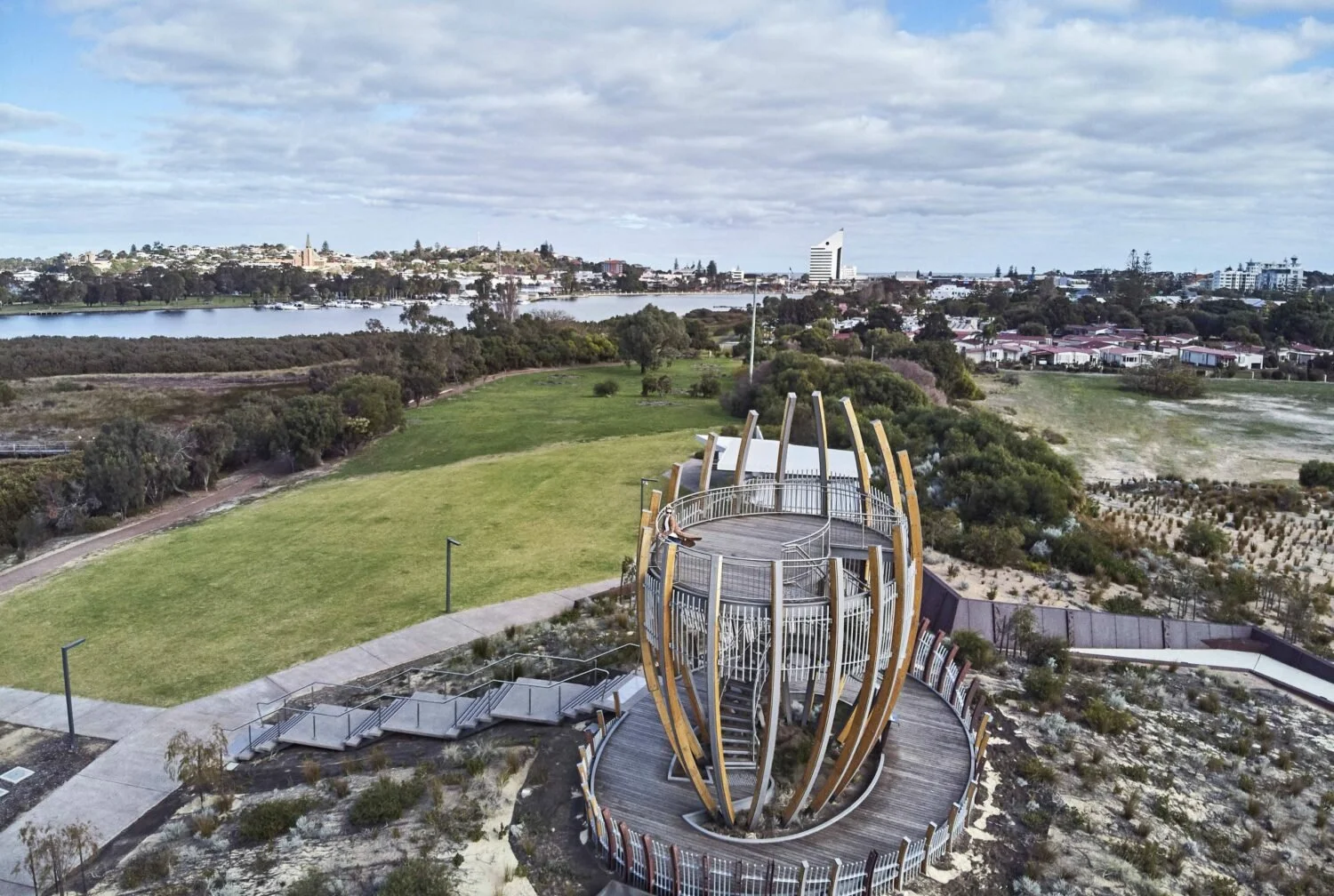 Aerial view of a park with a modern, spherical wooden structure, green grass, walking paths, and cityscape in the background, including water and buildings under a partly cloudy sky.