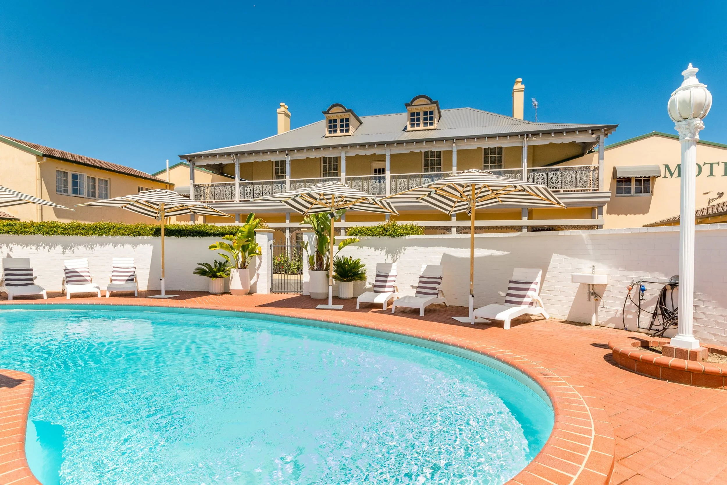 Swimming pool with white lounge chairs and striped umbrellas, surrounded by plants and a white brick wall, with a large two-story building with a balcony in the background under a clear blue sky.