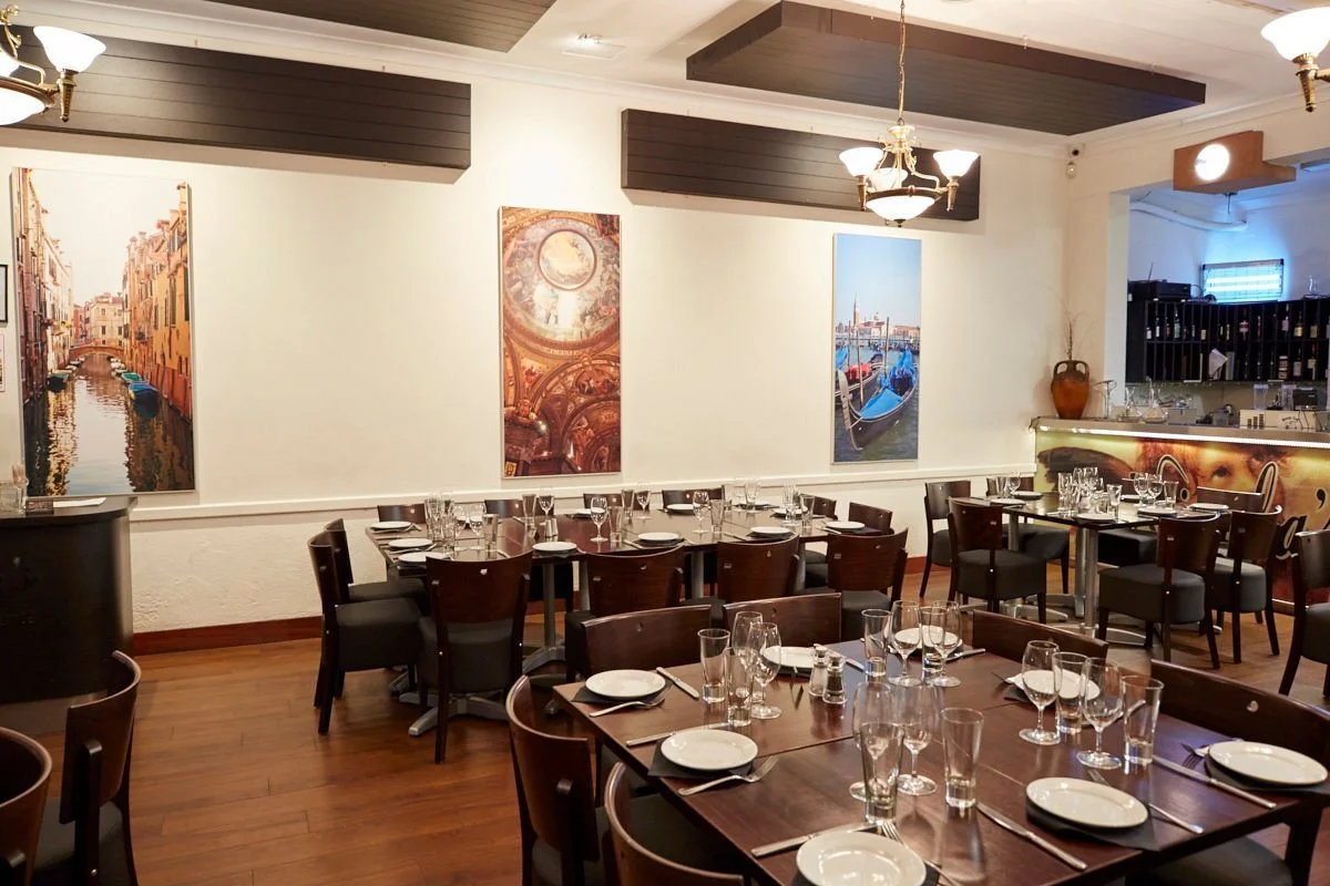 A cozy restaurant with wooden tables set with plates, glasses, and utensils, decorated with colorful artworks of Venice scenes on the white walls.