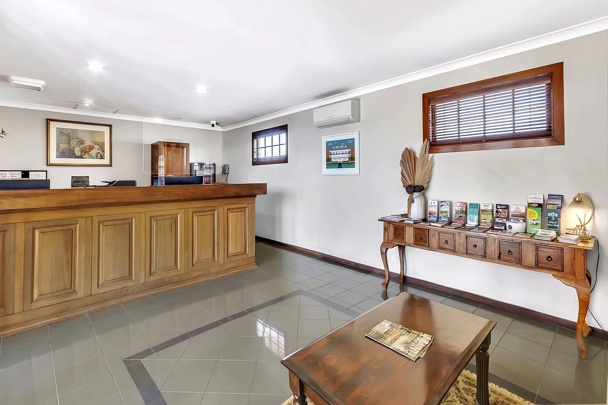 Hotel lobby with wooden reception desk, small wooden table, and decorative console with brochures and a lamp, two small windows with blinds, framed artwork on wall, air conditioning unit, and light-colored tiled floor.