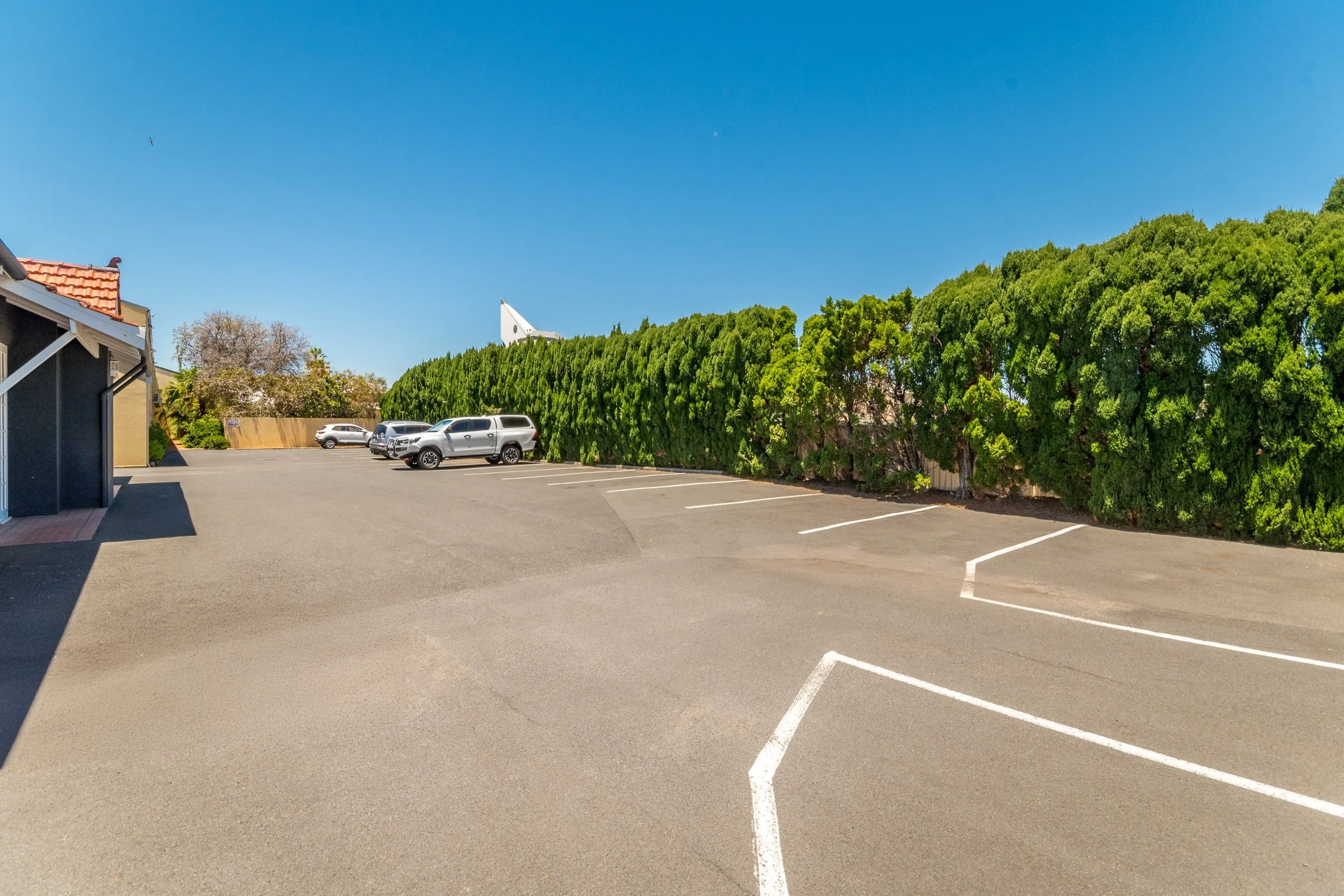 Empty parking lot with a few parked cars, a row of tall green bushes, and a small building with a red-tiled roof on the left, under a clear blue sky.