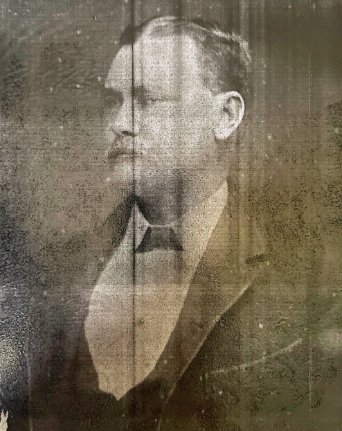 Black and white portrait of a man in formal attire with glasses, looking to the left, with a blurred background.