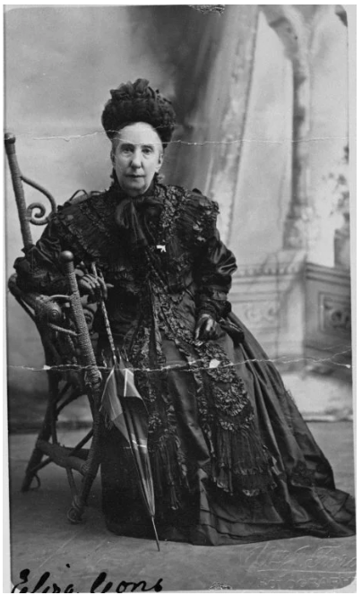 A black and white photo of a woman dressed in Victorian-era clothing, sitting on a wooden chair. She has her hair styled up and is wearing a high-collared dress with lace and intricate detailing. There is a parasol resting beside her, and the background features a painted backdrop with architectural elements.