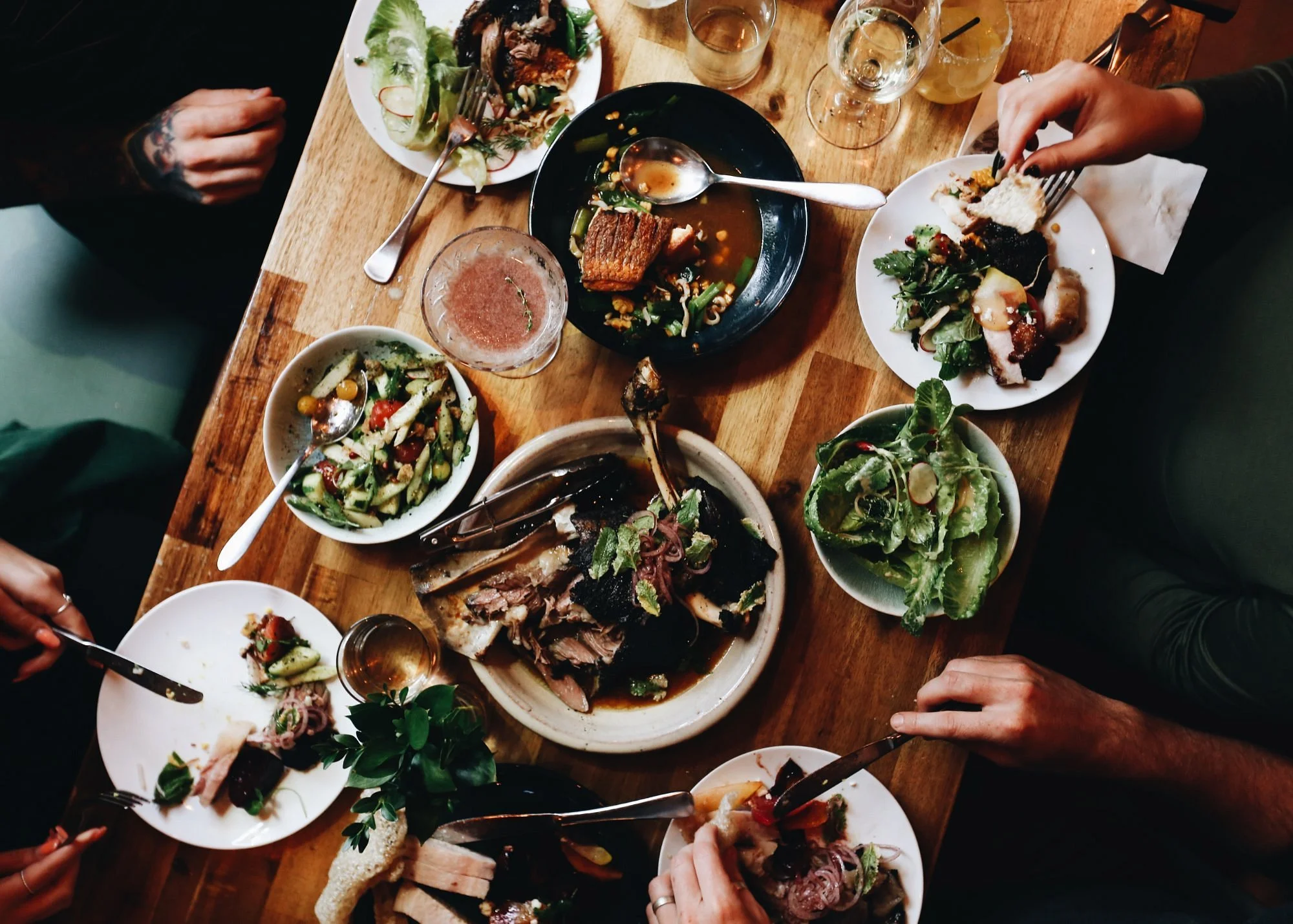 A wooden table filled with various dishes, including salads, roasted meat, and small dishes, with multiple hands reaching for food and drinks.