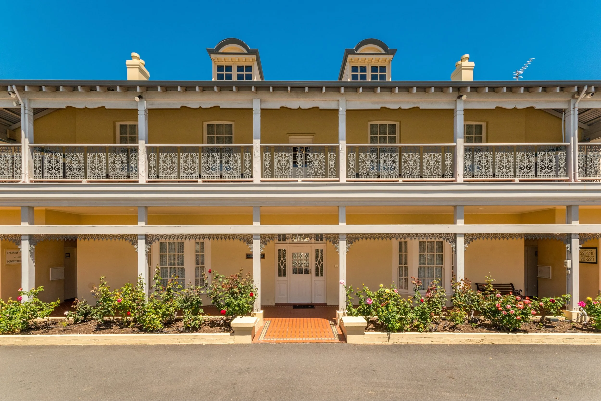 Front view of a yellow multi-story building with decorative white balcony railings and flower beds with pink and red roses, under a clear blue sky.