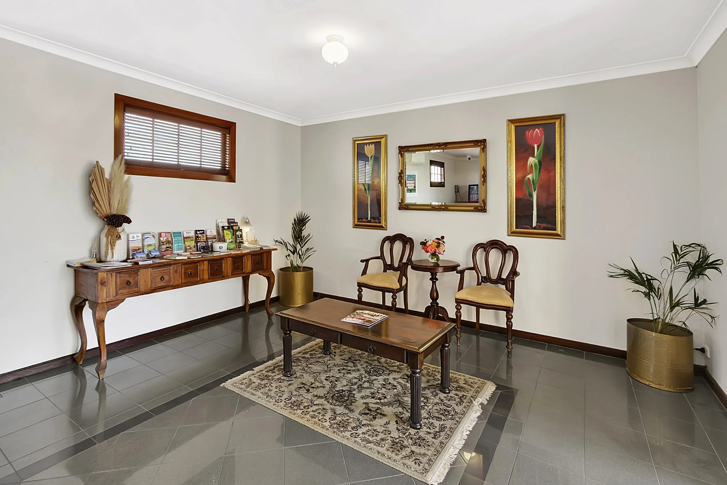 Hotel lobby with a wooden table, two chairs with beige cushions, a mirror, framed flower artworks, potted plants, and a display table with brochures on the wall.