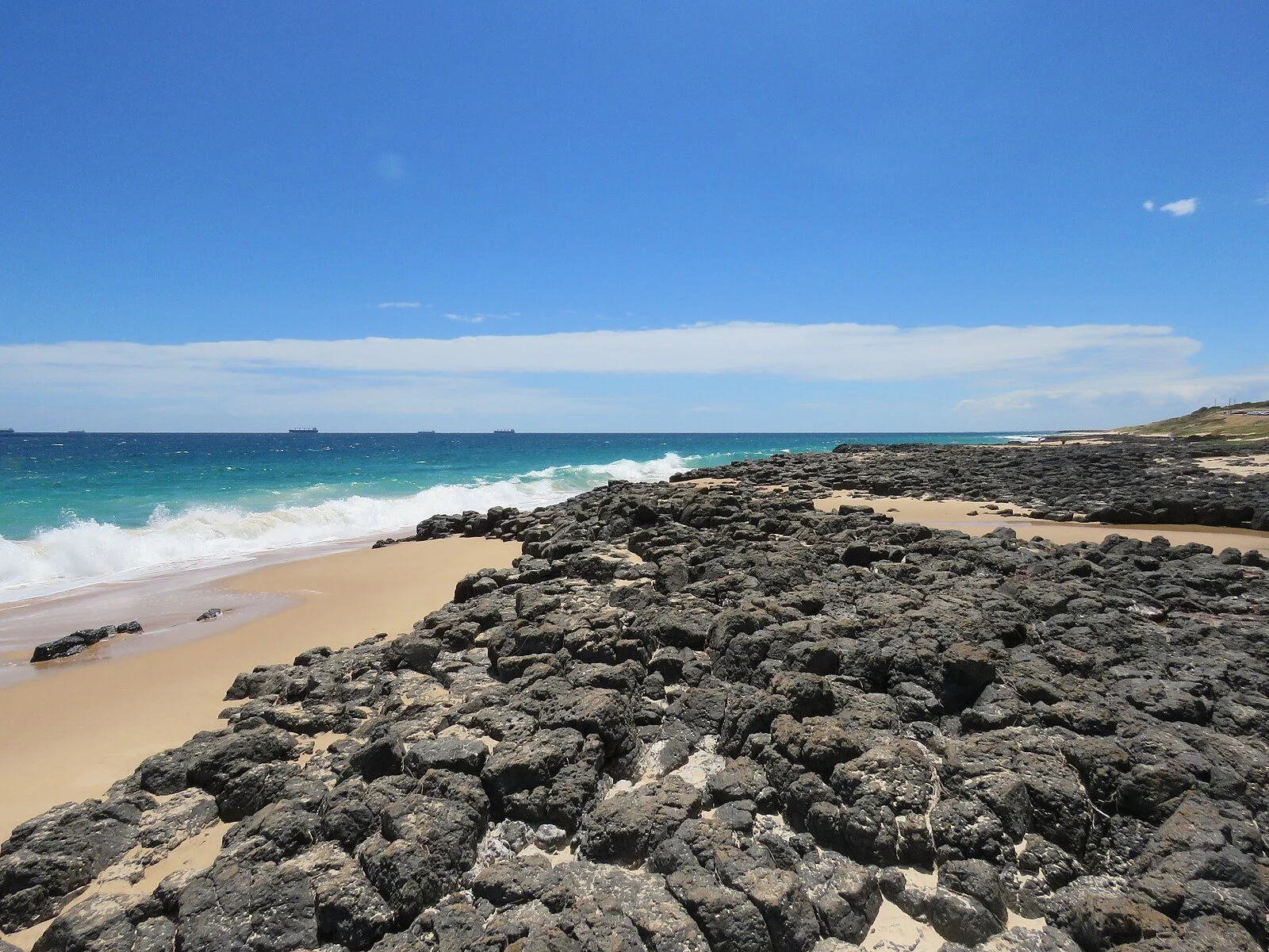 A rocky beach with dark rocks along a sandy shore, turquoise ocean waves, and a clear blue sky with some clouds.