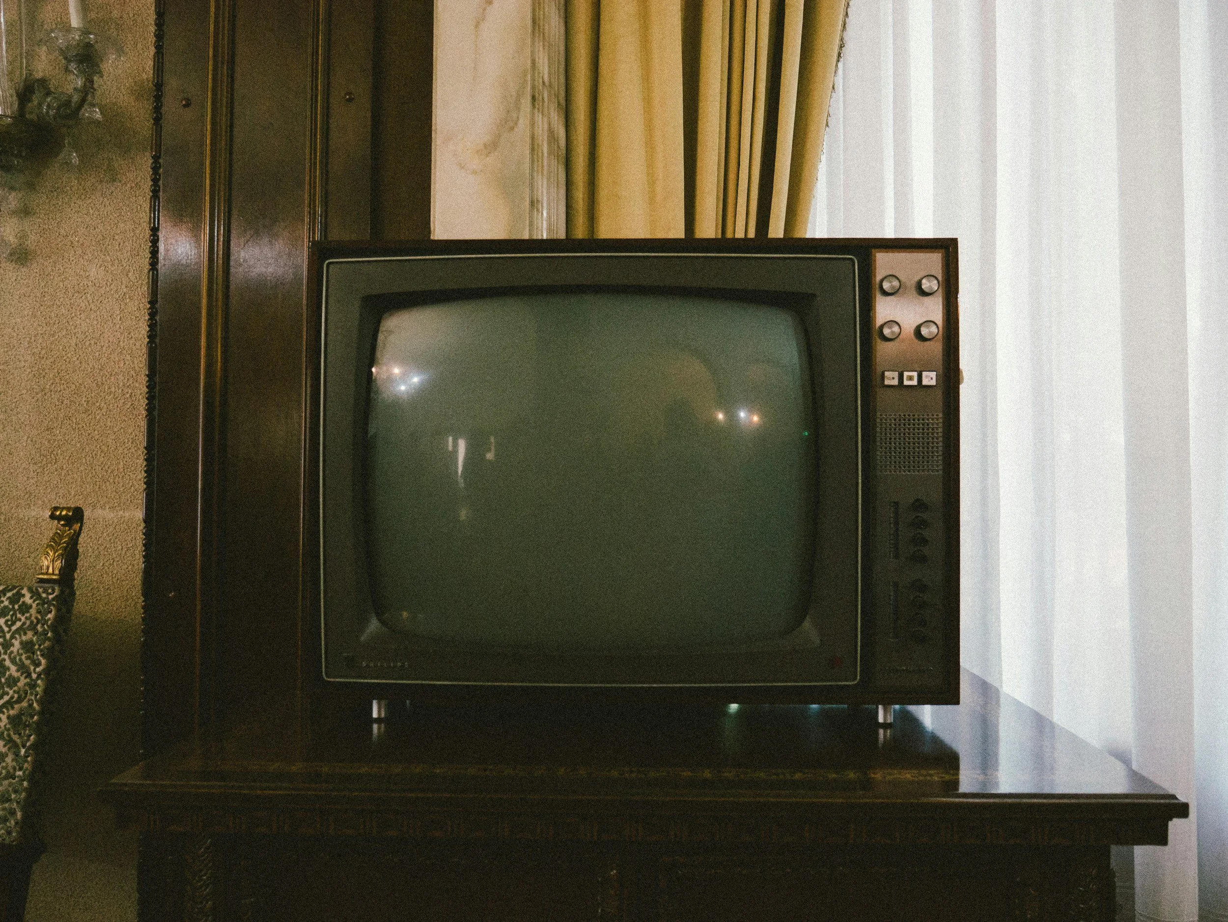 An old CRT television on a dark wooden table in a vintage living room with beige walls, curtain, and gold-patterned chair.