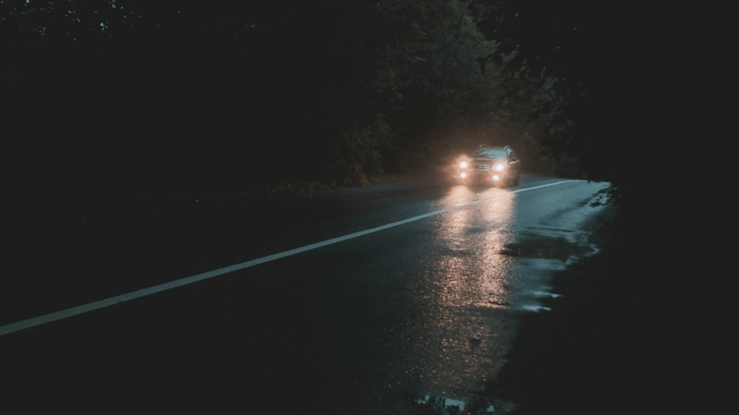 A car driving on a wet, dark road at night with headlights on, surrounded by trees.