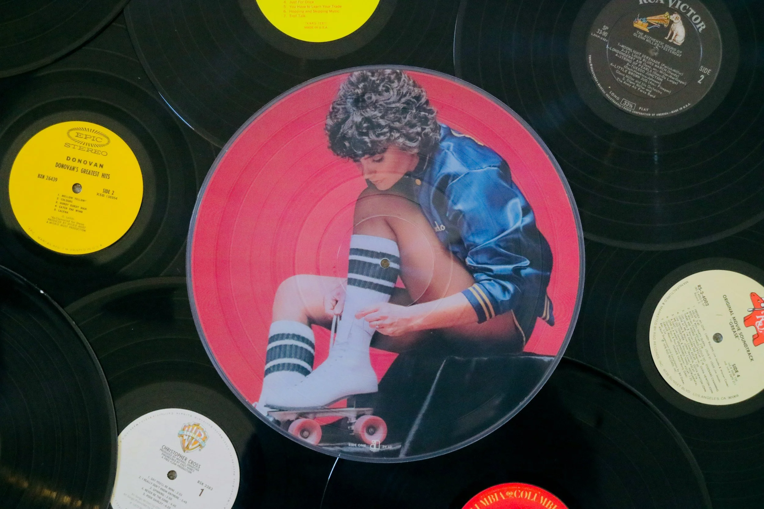 A collection of vinyl records with a central record featuring a woman in a blue jacket and striped socks, tying her shoelace while sitting on a skateboard.