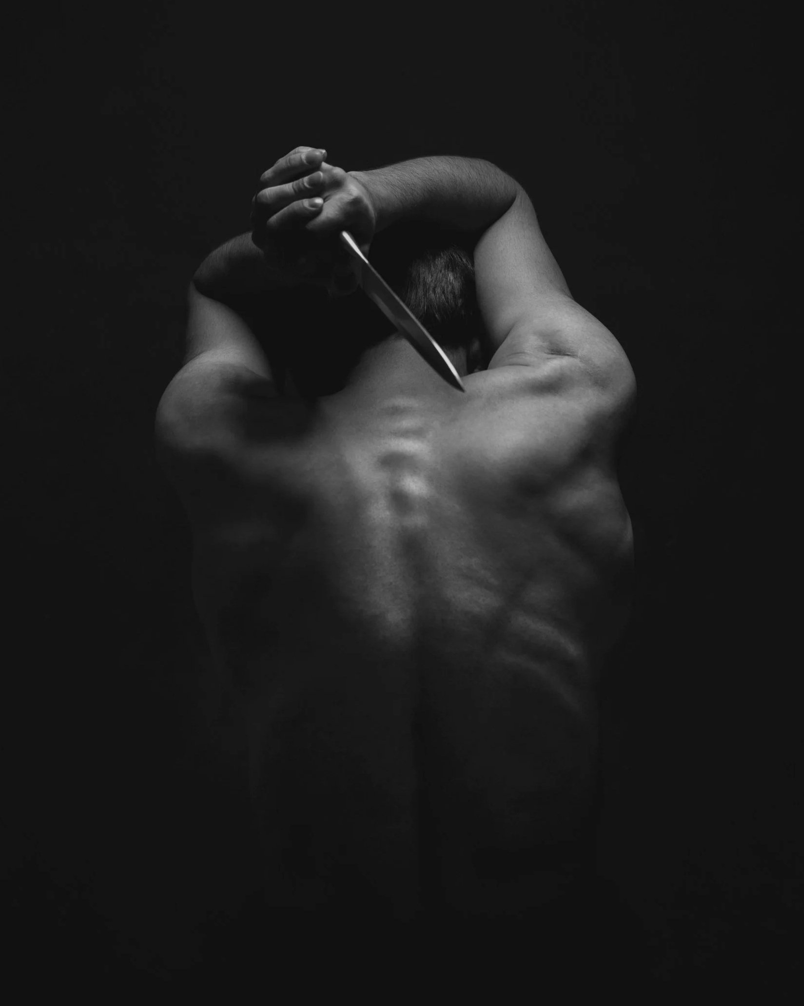 A black-and-white photo of a shirtless man with his back to the camera, holding a knife above his head with one hand and the other hand around his head, against a dark background.