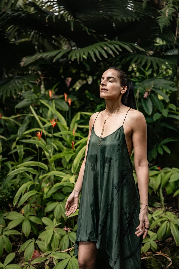 A person standing with a green dress surrounded by rich botanical textures while closing their eyes in peace