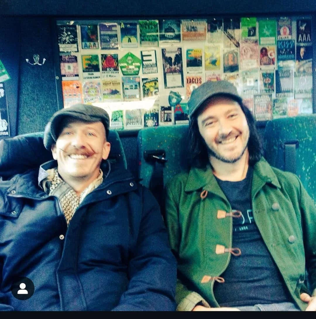 Two smiling men sitting on a bus or train with a wall of posters behind them.