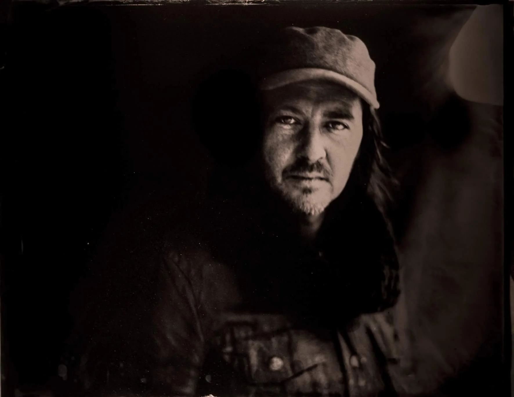 Black and white portrait of a man wearing a cap and a leather jacket, with long hair, looking into the camera with a serious expression.