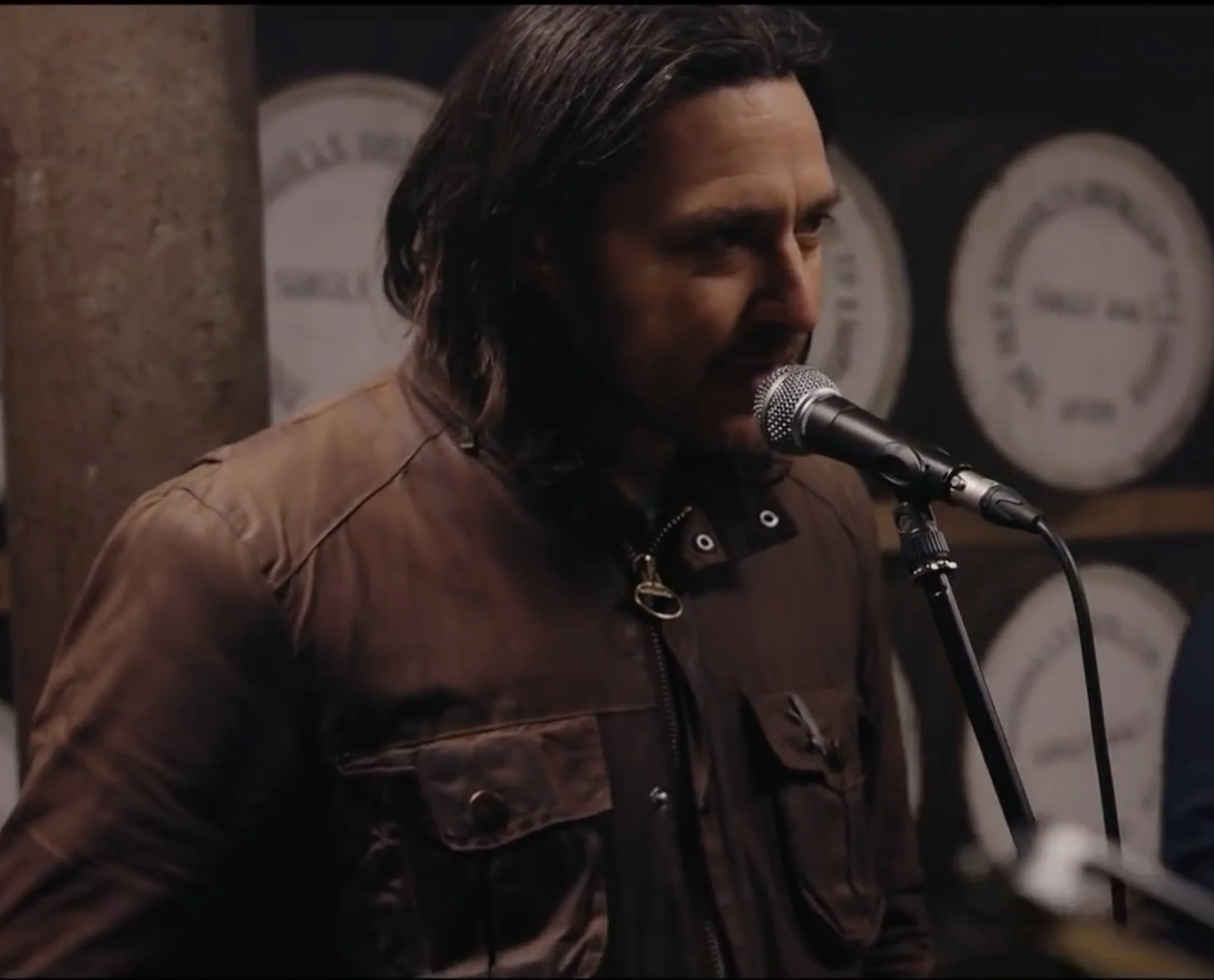 A man singing into a microphone in a dimly lit room with gauges on the wall in the background.
