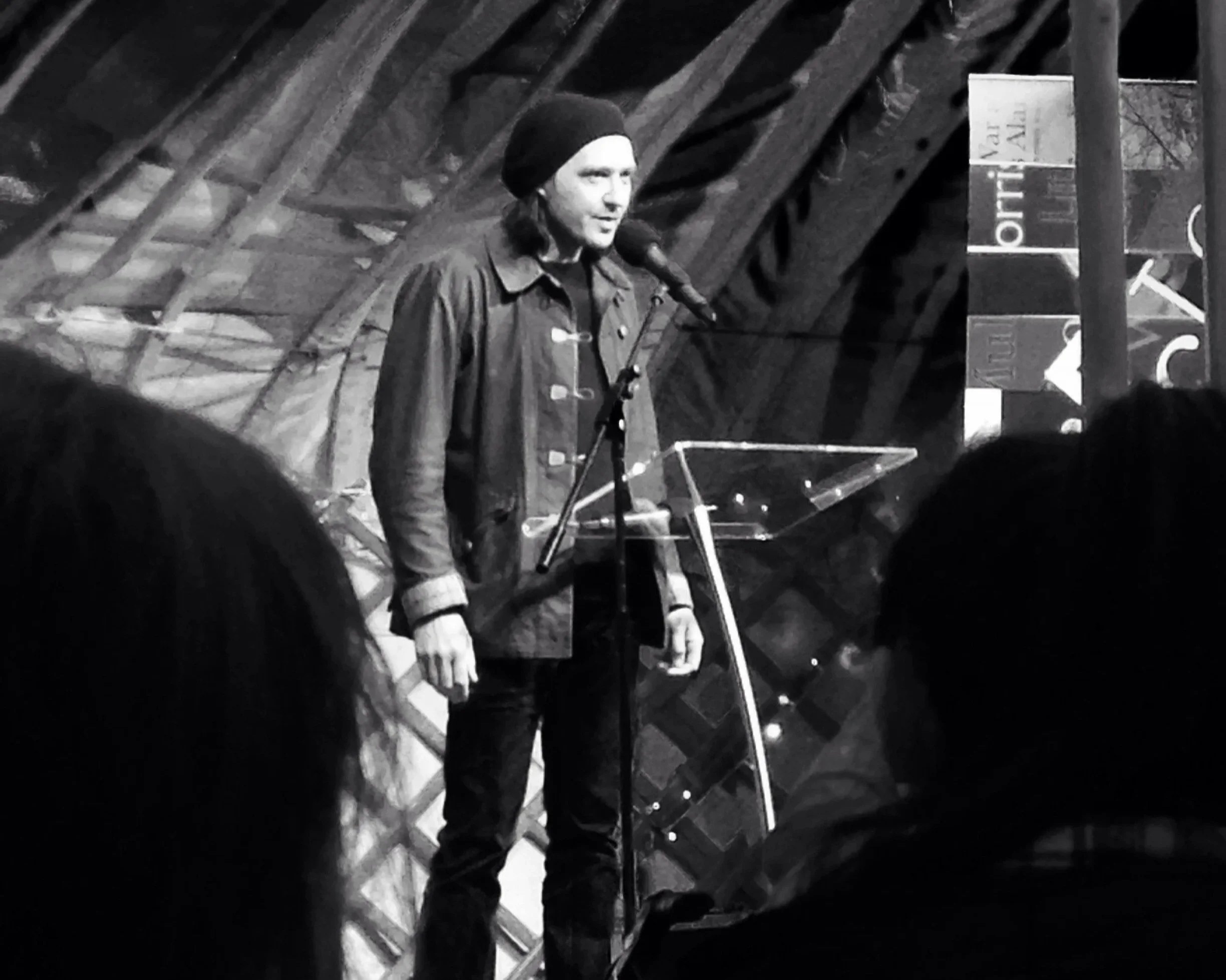 A person (Jon Plunkett) is standing at a microphone on a stage, wearing a black beanie and a black jacket, with a music stand in front of them, in front of a background of wooden beams.