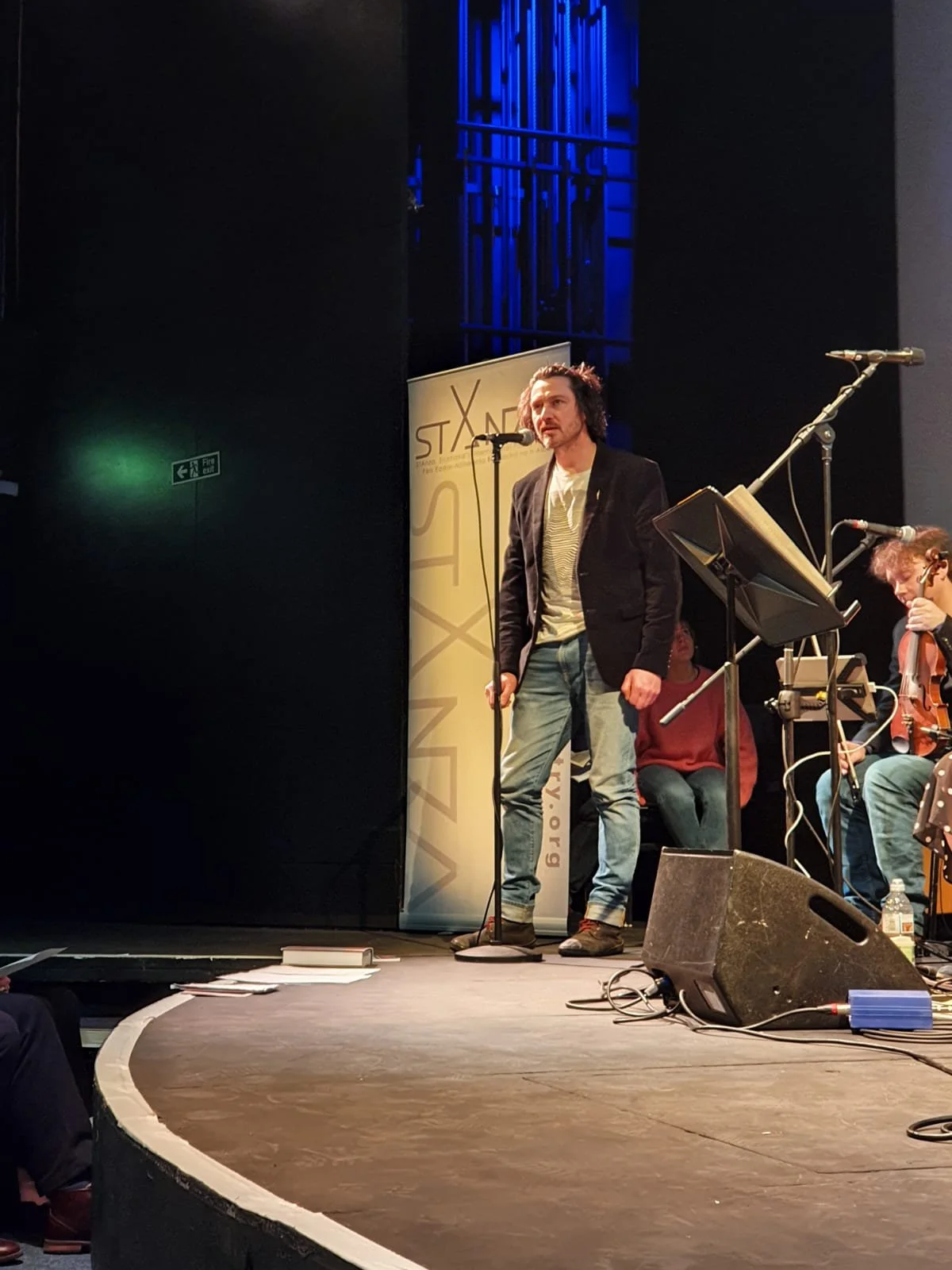 A man with curly hair and facial hair stands on a stage, speaking or singing into a microphone. Behind him, a band with a violinist and a person in a red shirt are visible. The stage has a black background with a vertical banner and technical equipment.
