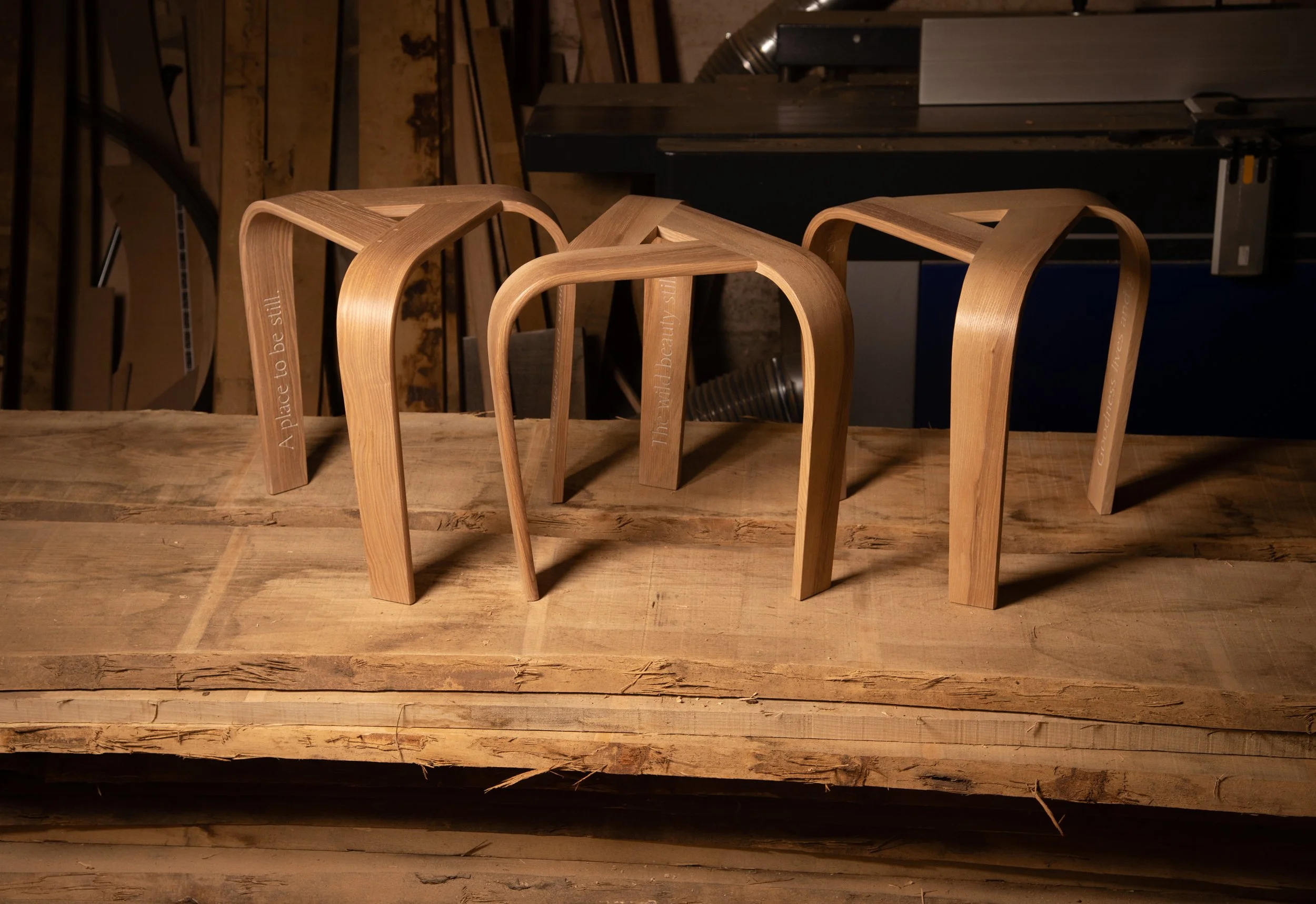 Three wooden stools on a rustic wooden table in a woodworking workshop made by Angus Ross and featuring lines of poetry on each leg by Jon Plunkett.