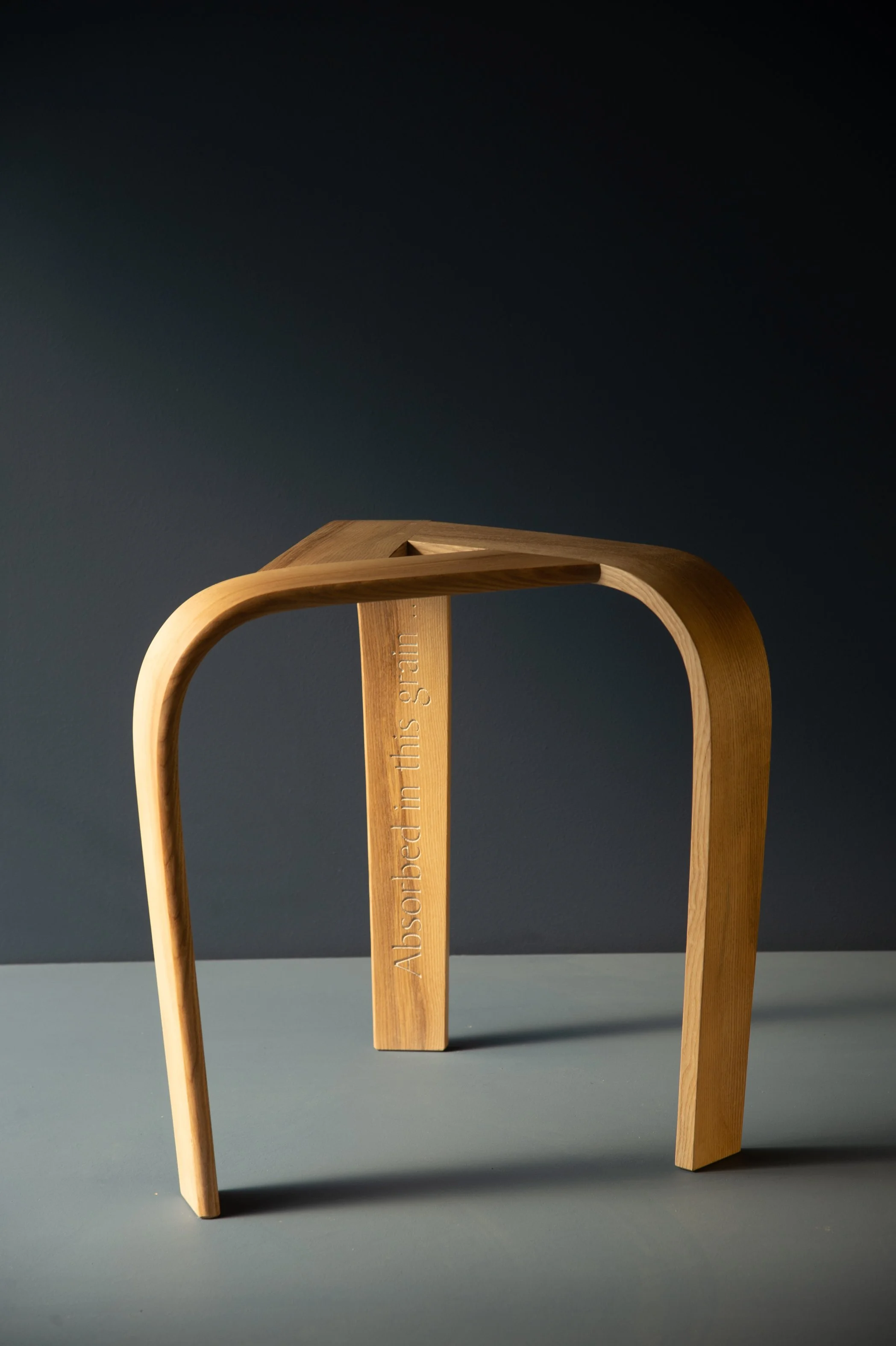 Modern wooden stool with an engraved phrase 'Absorbed in this grain' on one leg, written by Jon Plunkett placed on a two-tone surface with a dark background.