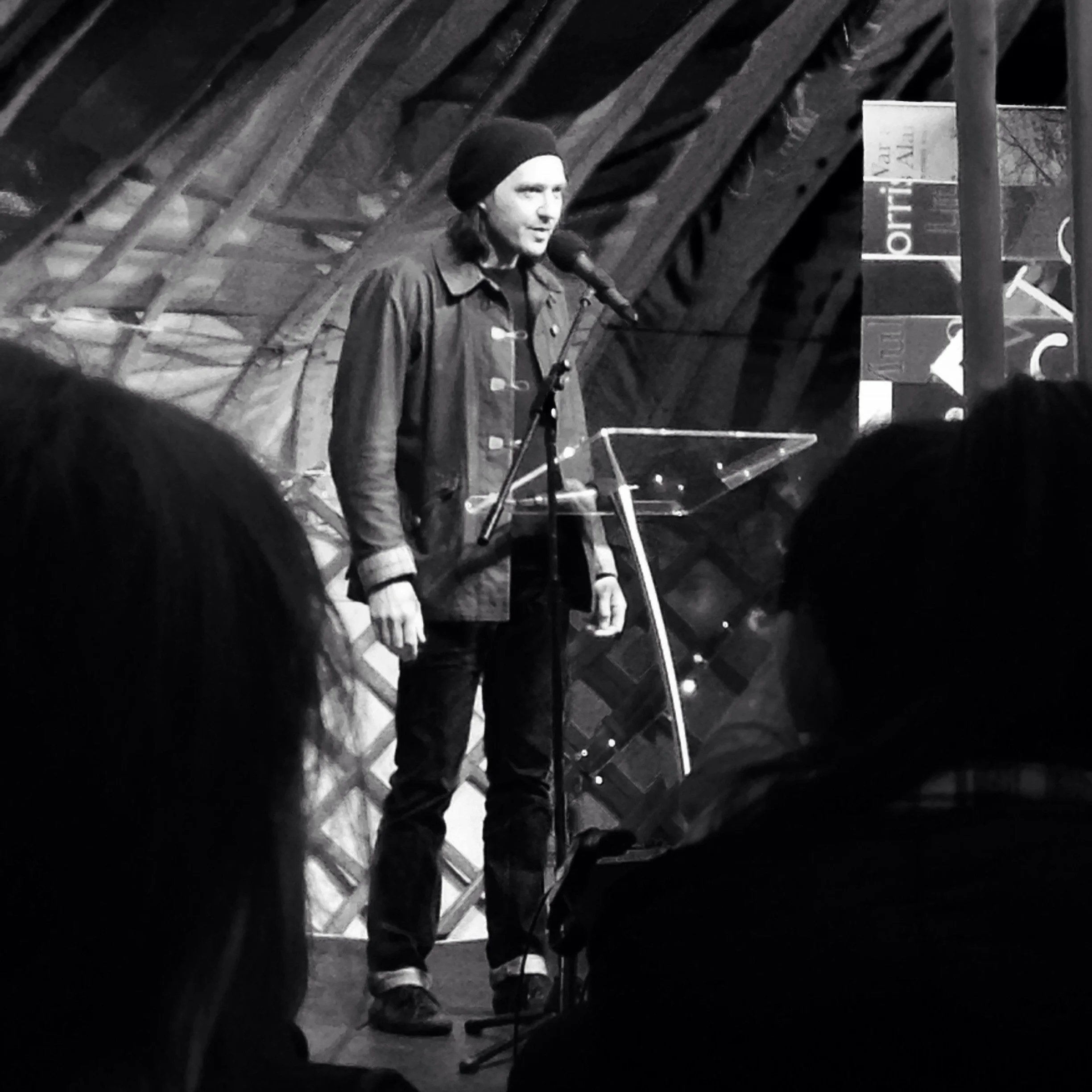 A person wearing a beanie hat and a jacket speaking into a microphone on a stage, with an audience in front.