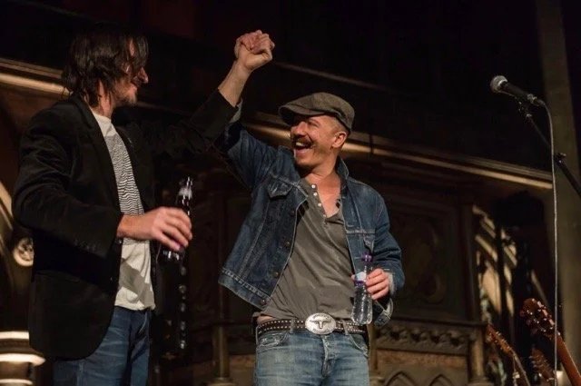 Two men on stage, one wearing a black jacket and the other wearing a denim jacket and cap, greeting each other with a fist bump, while holding drinks.