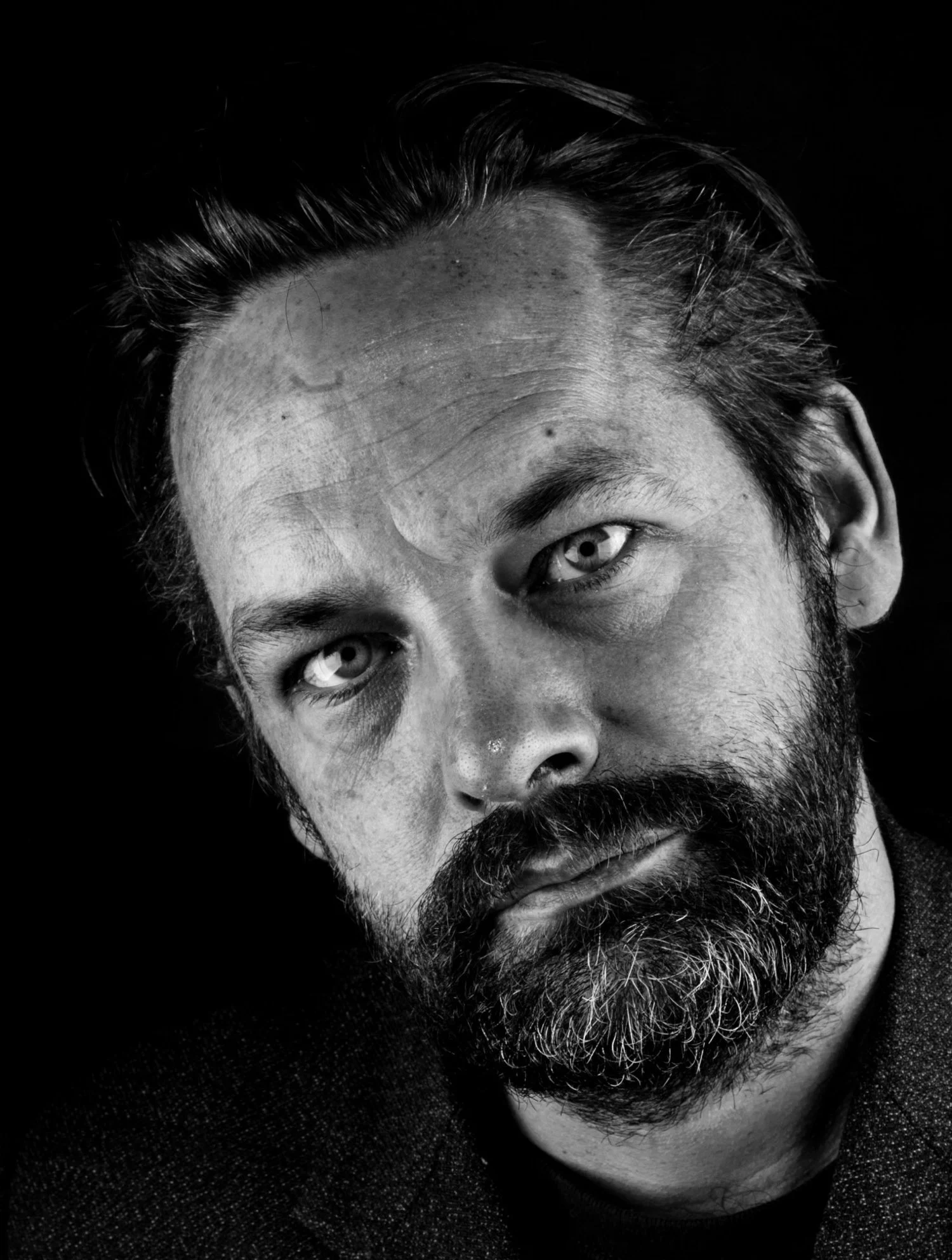 Black and white self-portrait of James Millar, intense gaze, and slightly disheveled hair, against a dark background.