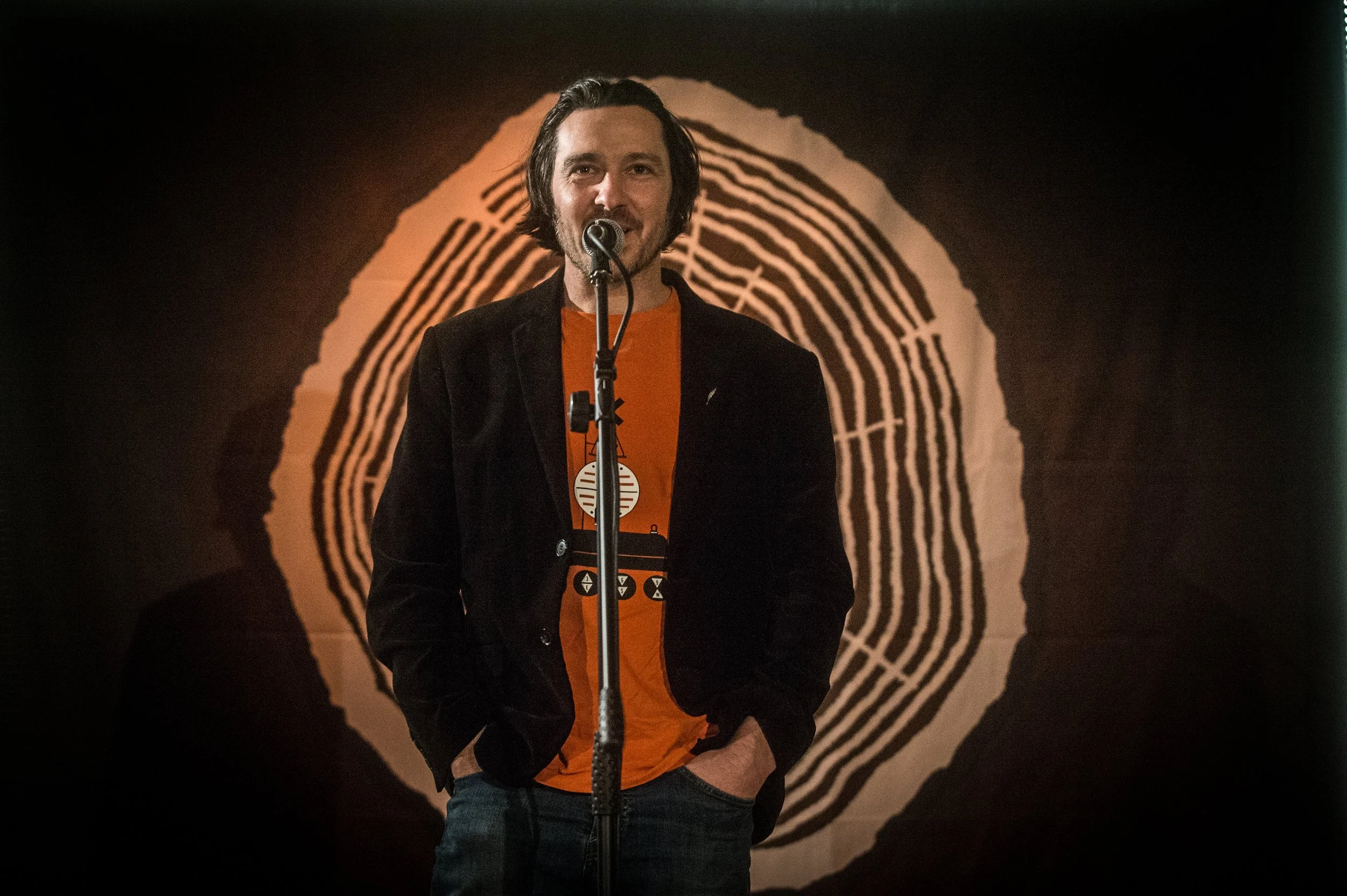 A man speaking into a microphone on stage, with a large fingerprint image as the backdrop.