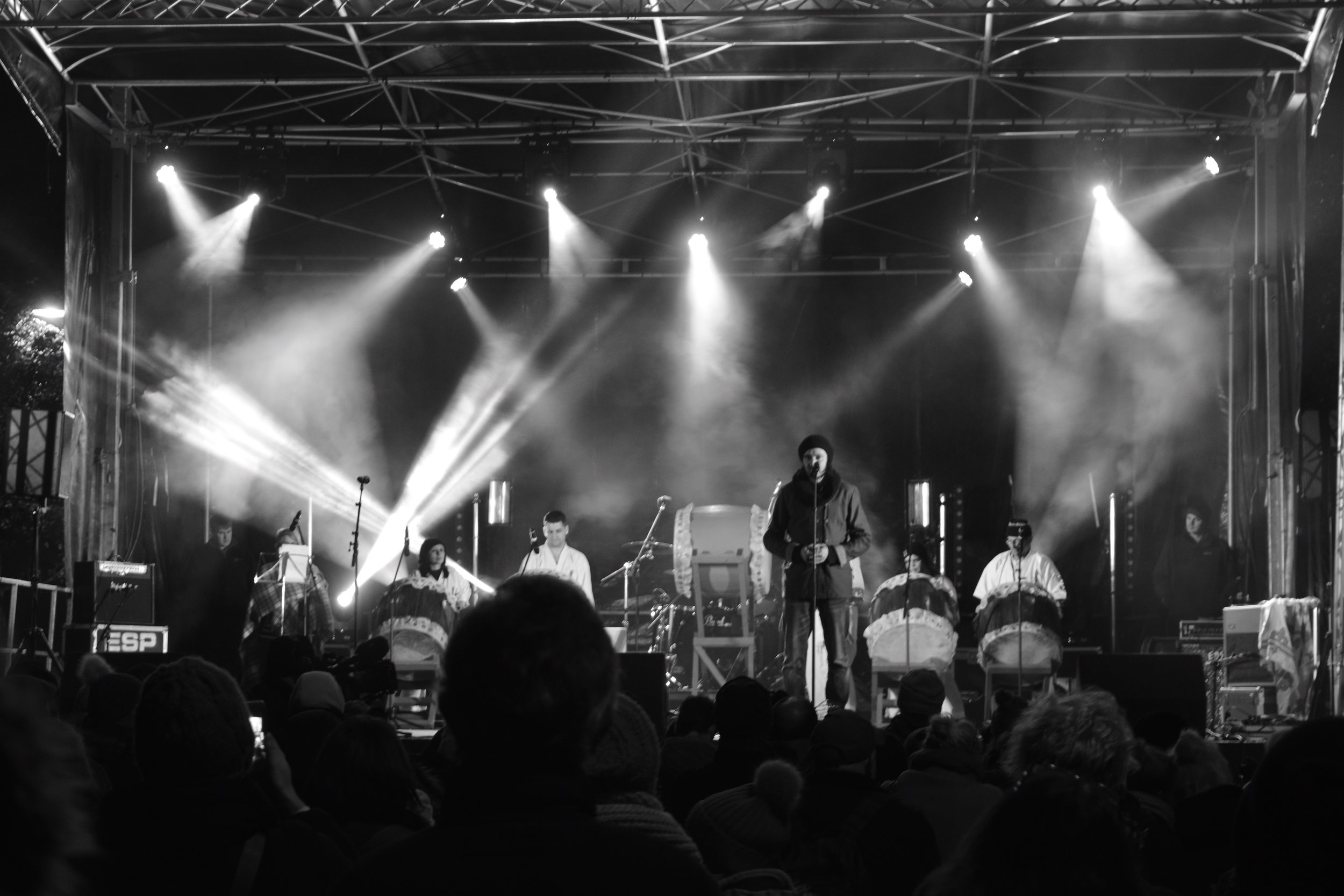 A black and white photo of a live outdoor music performance on a stage with a group of musicians, some playing drums and other instruments, and a singer at the front. Audience members are visible in the foreground, watching the performance.